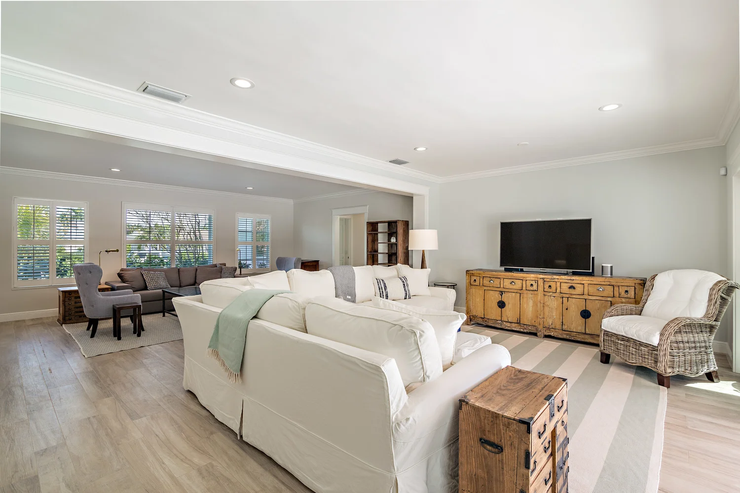 Bright living room with white sofas, wicker chair, wooden console, TV, large windows with blinds, and light-colored hardwood floors.