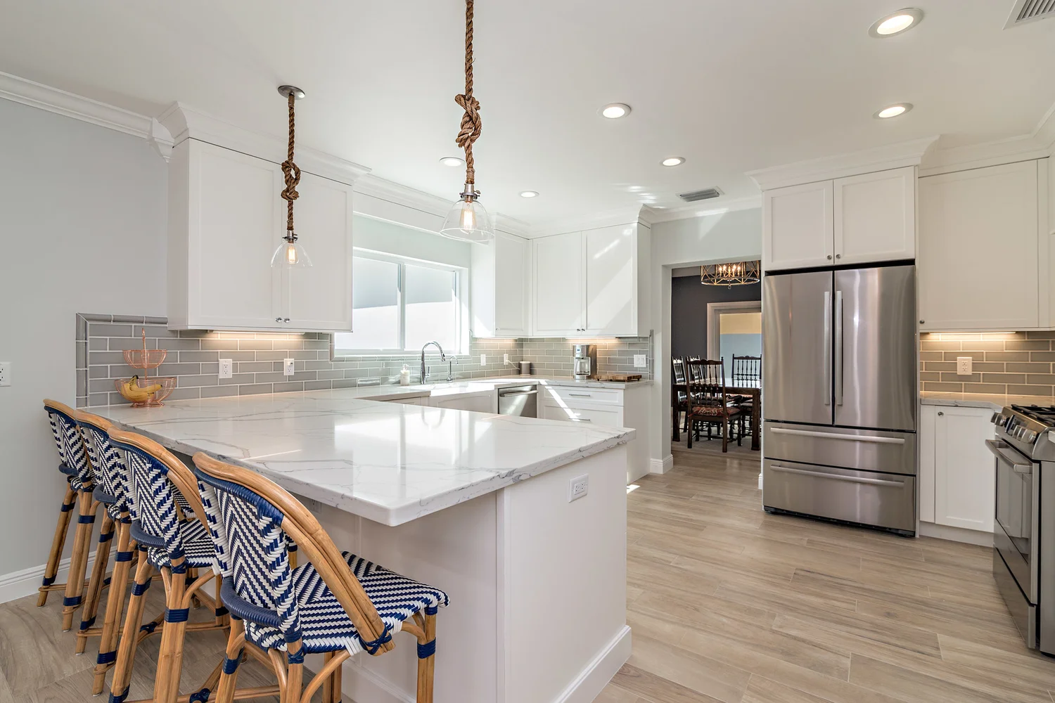 Modern kitchen with white cabinets, gray subway tile backsplash, marble island with seating, and stainless steel appliances, illuminated by recessed lighting and two hanging pendant lights with rope cords.