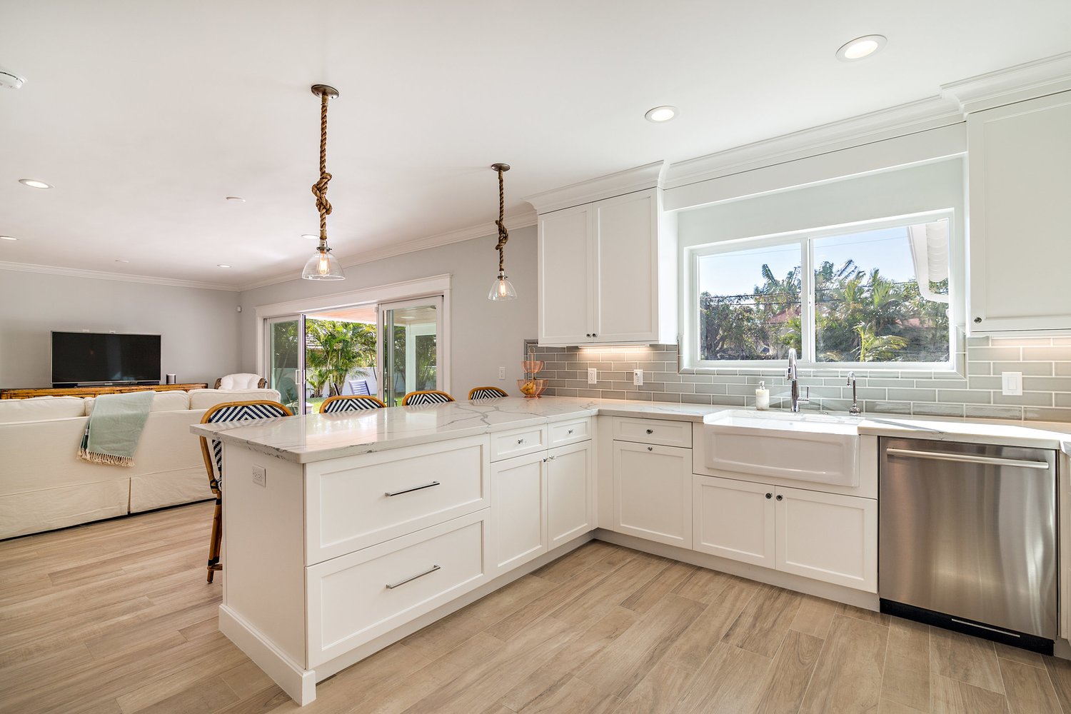 Modern kitchen with white cabinets, a marble countertop island, large window overlooking greenery, and hanging pendant lights with rope details.