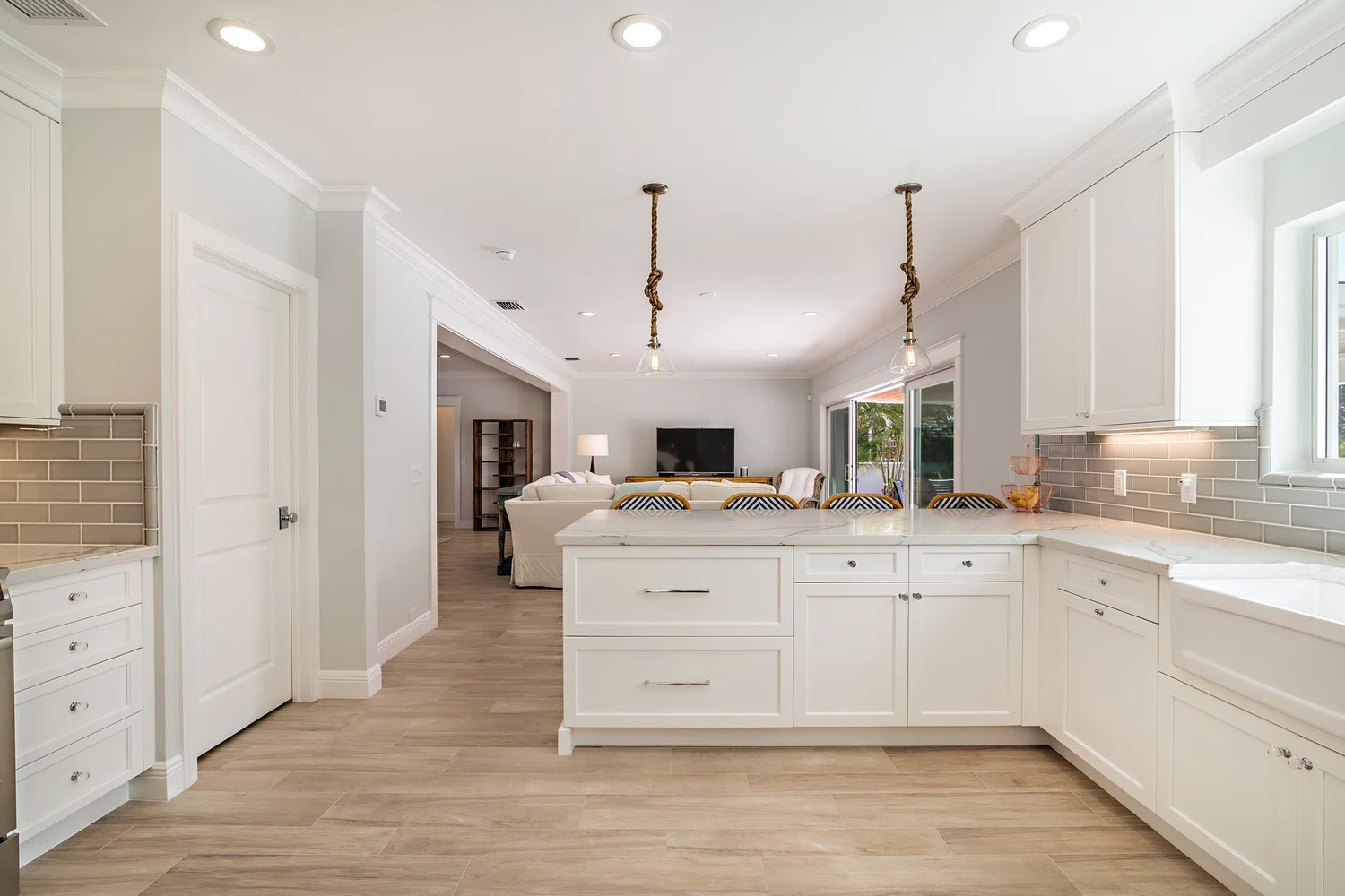 Interior view of a modern kitchen with white cabinets, a marble island, and wood flooring. The open-plan space extends into a living area with a television, sofa, and large windows.