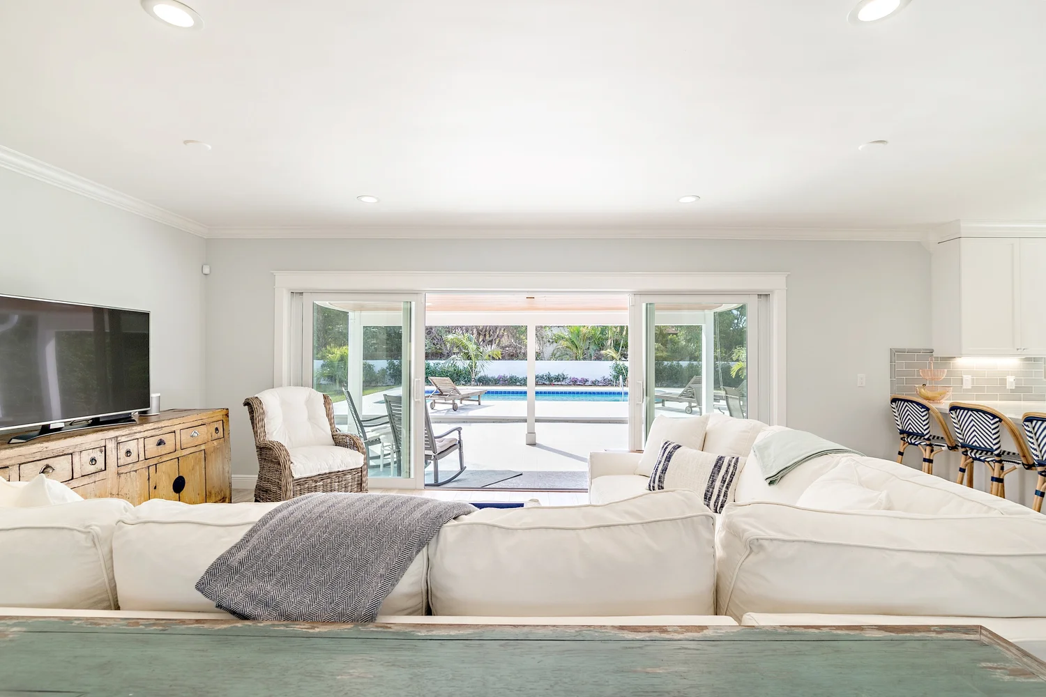 Living room with white sofas, a wooden cabinet with a TV, and large sliding glass doors leading to a patio with outdoor furniture and a pool, overlooking a backyard with trees.