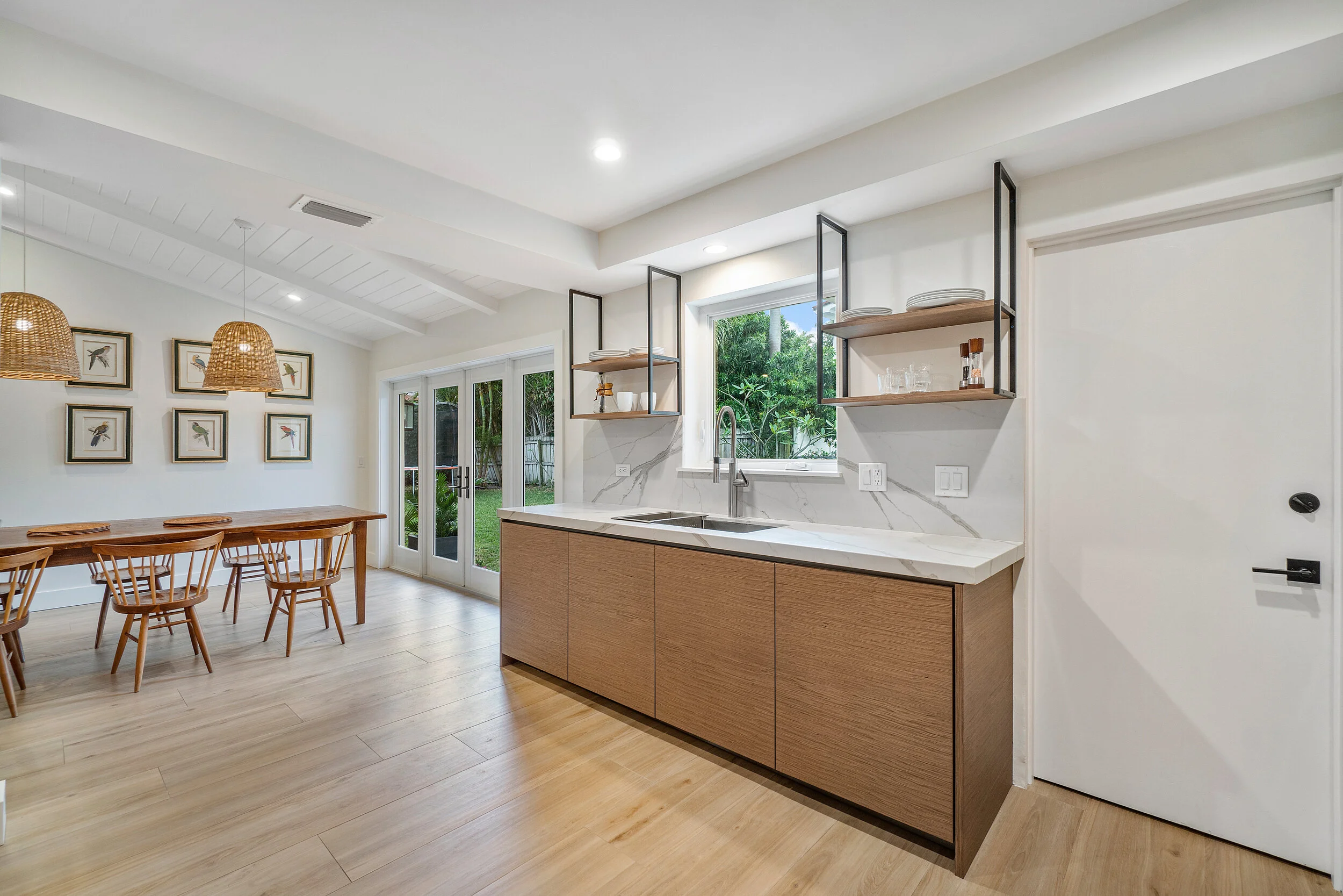 Modern kitchen and dining area with wood cabinetry, marble-look countertops, open shelving, woven pendant lights, and doors opening to the yard.