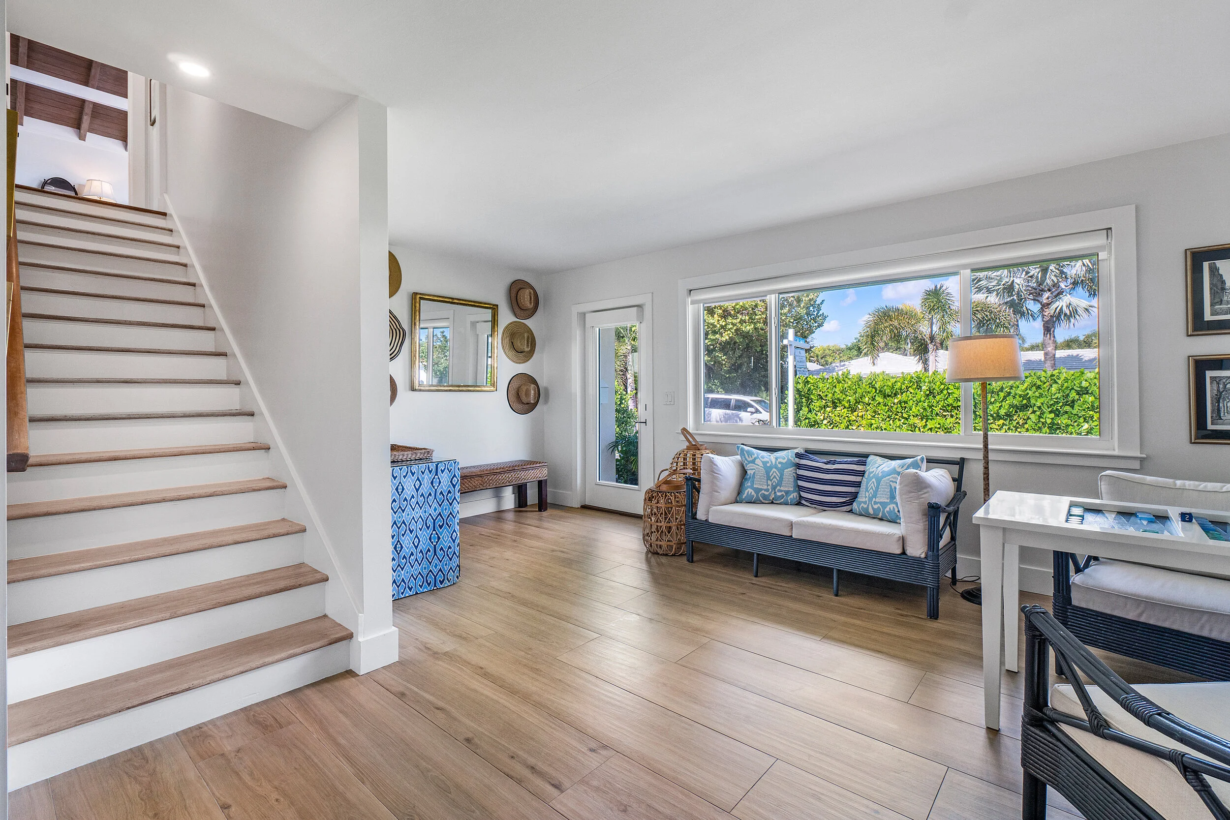 Light-filled sitting area with wood staircase, large window, blue and white sofa, game table, wood floors, and coastal-style decor.