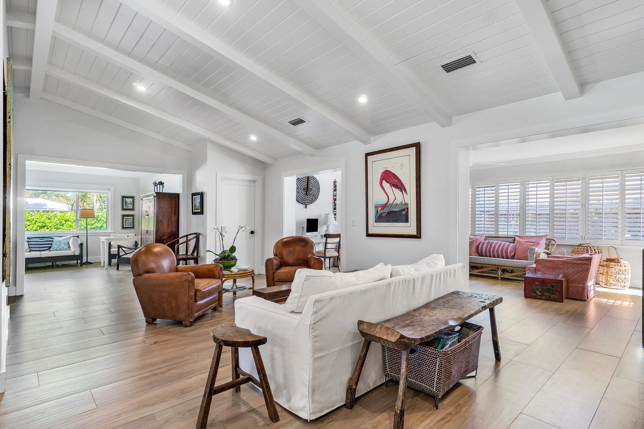 Spacious open living area with white sofa, leather chairs, vaulted ceiling beams, wood floors, framed flamingo artwork, and adjacent sunroom.