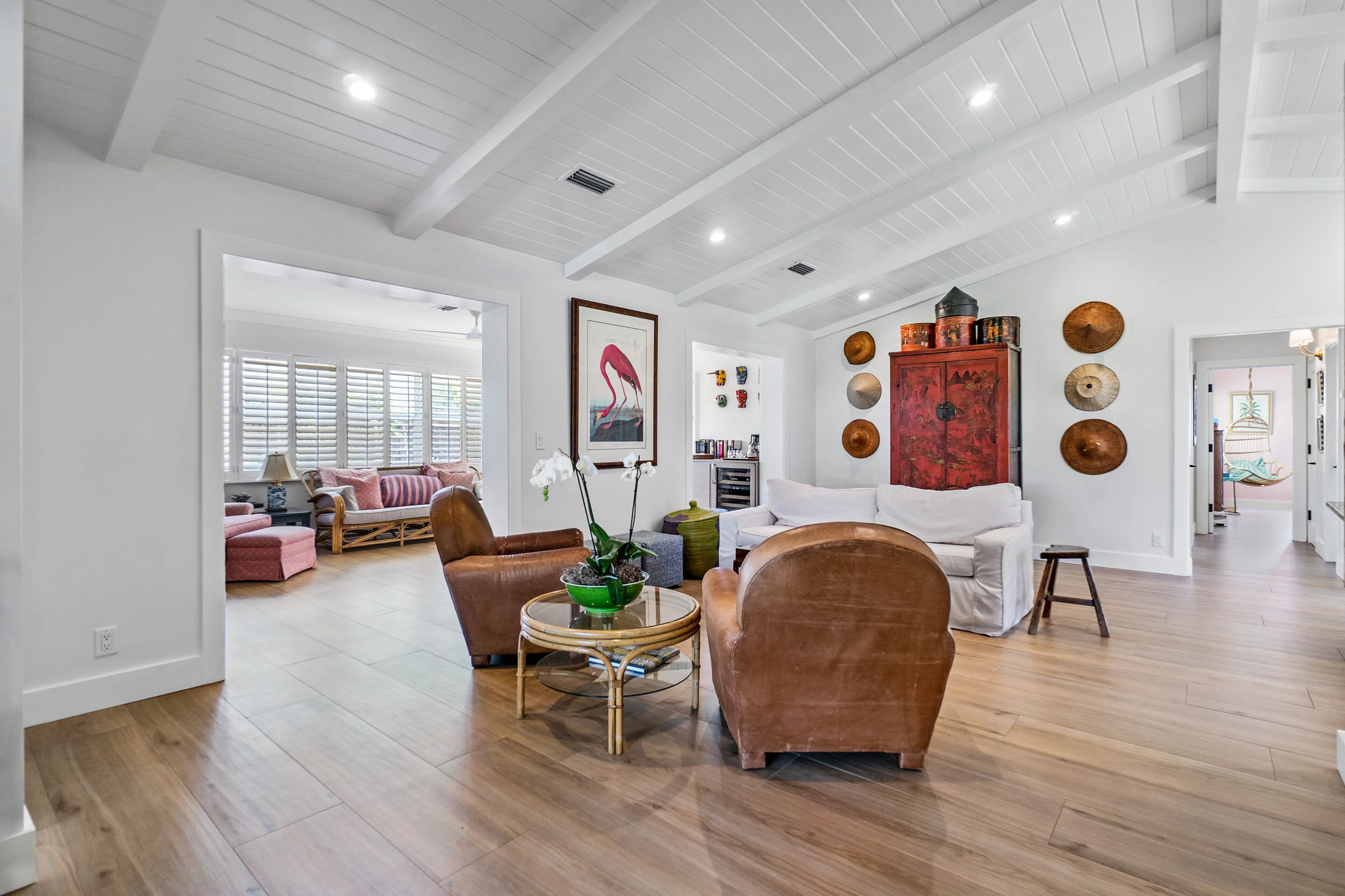 Bright living room with exposed white ceiling beams, leather armchairs, white sofa, wood floors, red cabinet, and adjoining sitting room.
