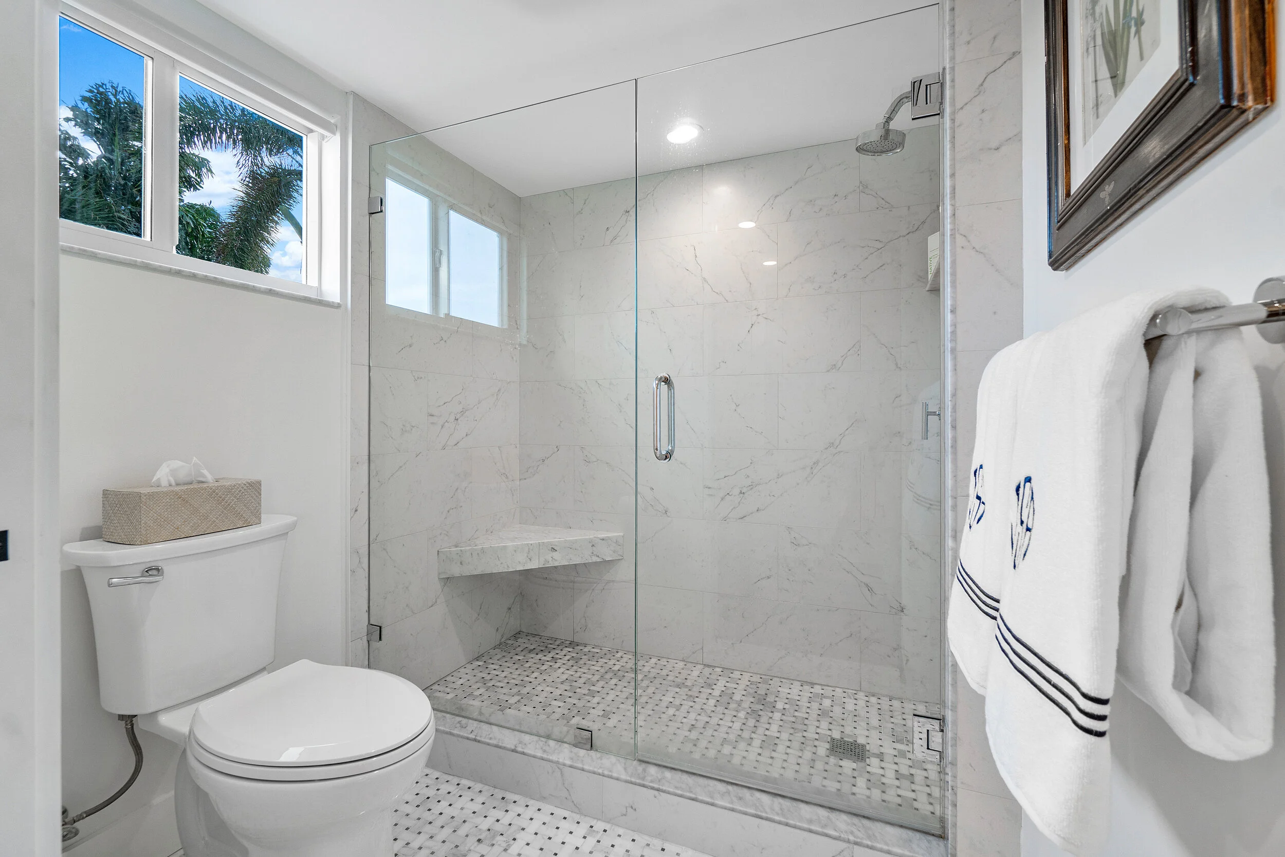 White bathroom with marble-look walk-in shower, glass enclosure, mosaic tile floor, small window, toilet, and folded towels on a wall rack.