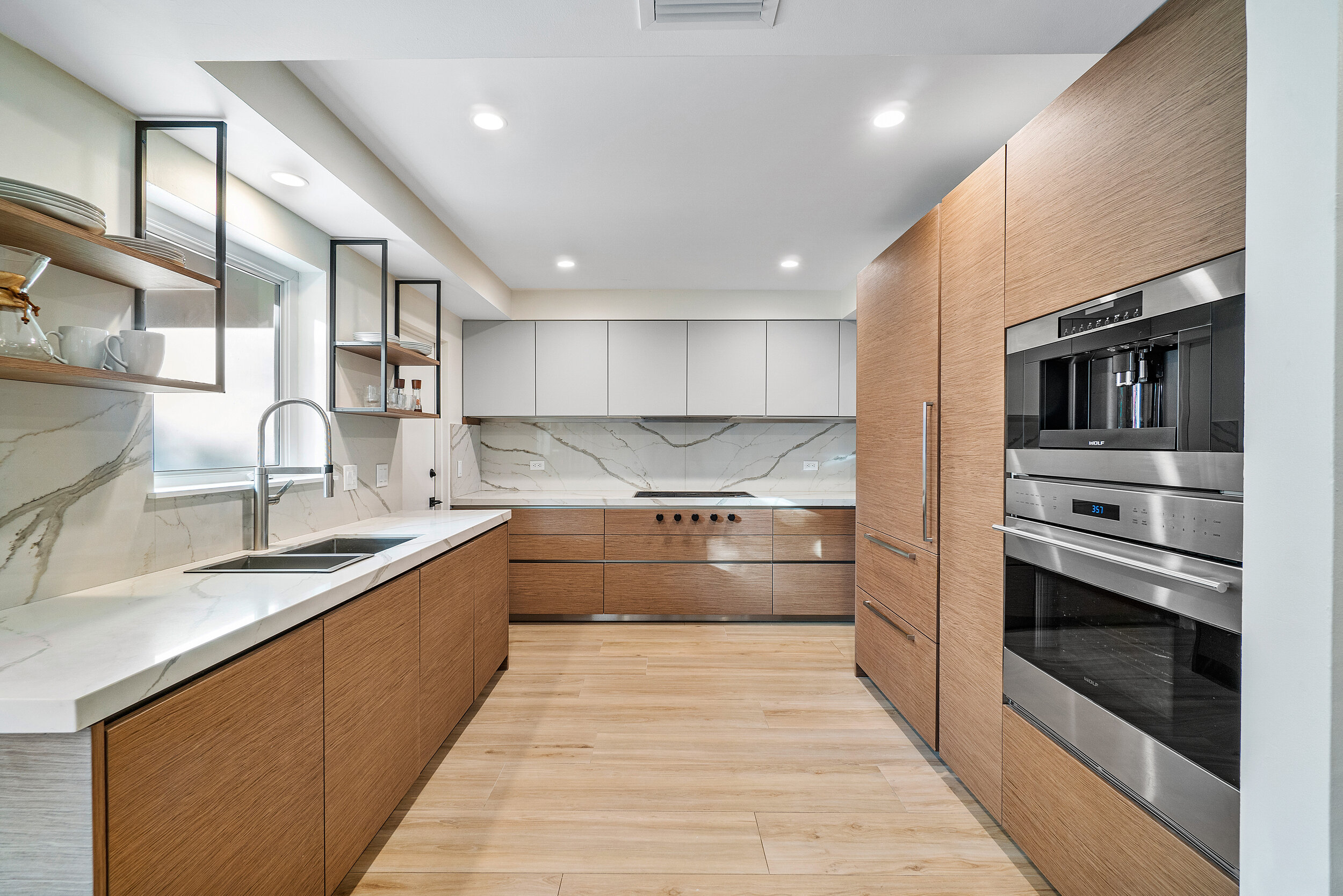 Contemporary kitchen with flat-panel wood cabinets, marble-look backsplash, stainless steel wall oven, built-in coffee machine, and open shelving.