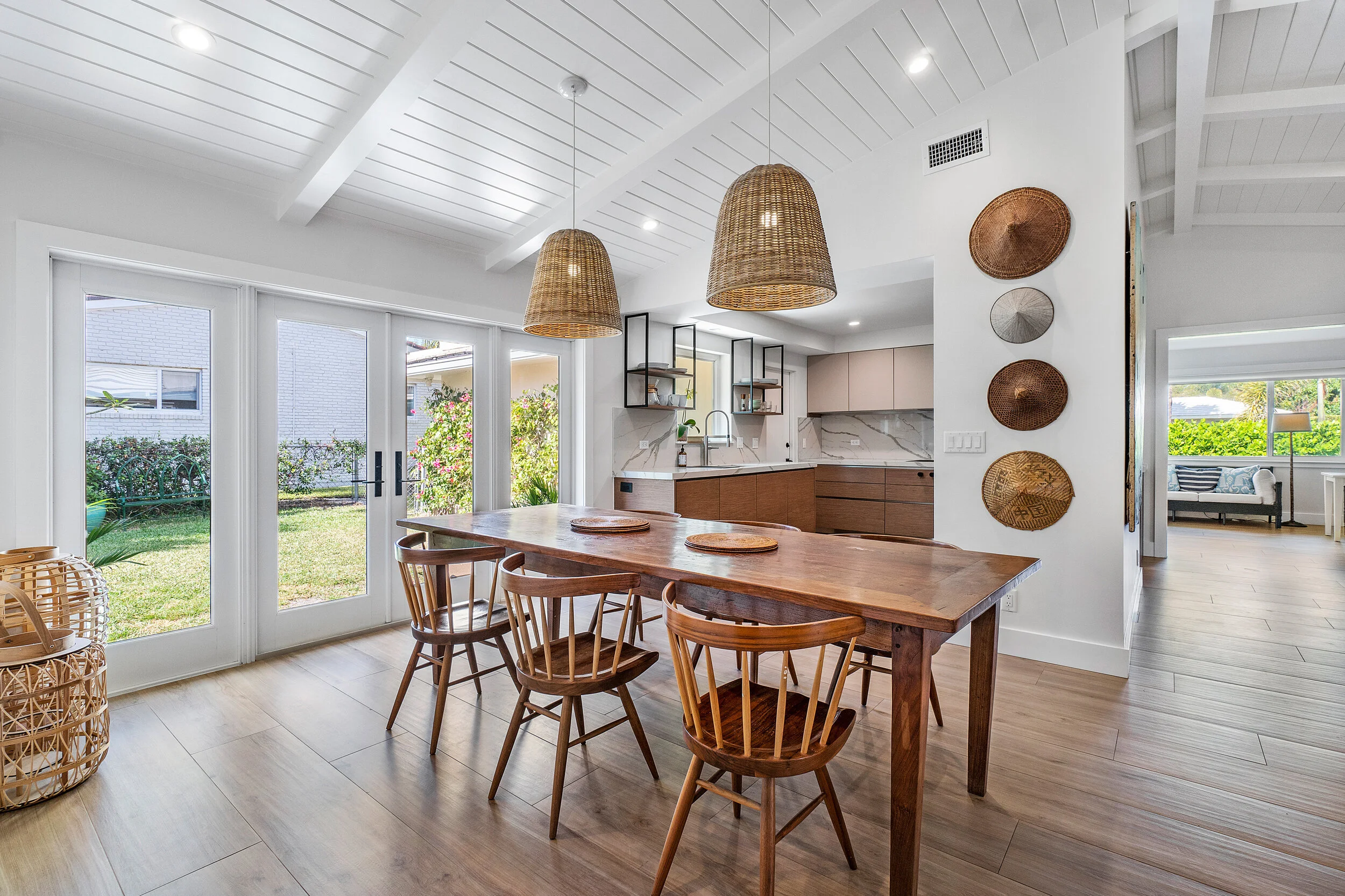 Open dining area with a wood table, woven pendant lights, glass doors to the yard, and a modern kitchen with wood cabinetry and marble backsplash.