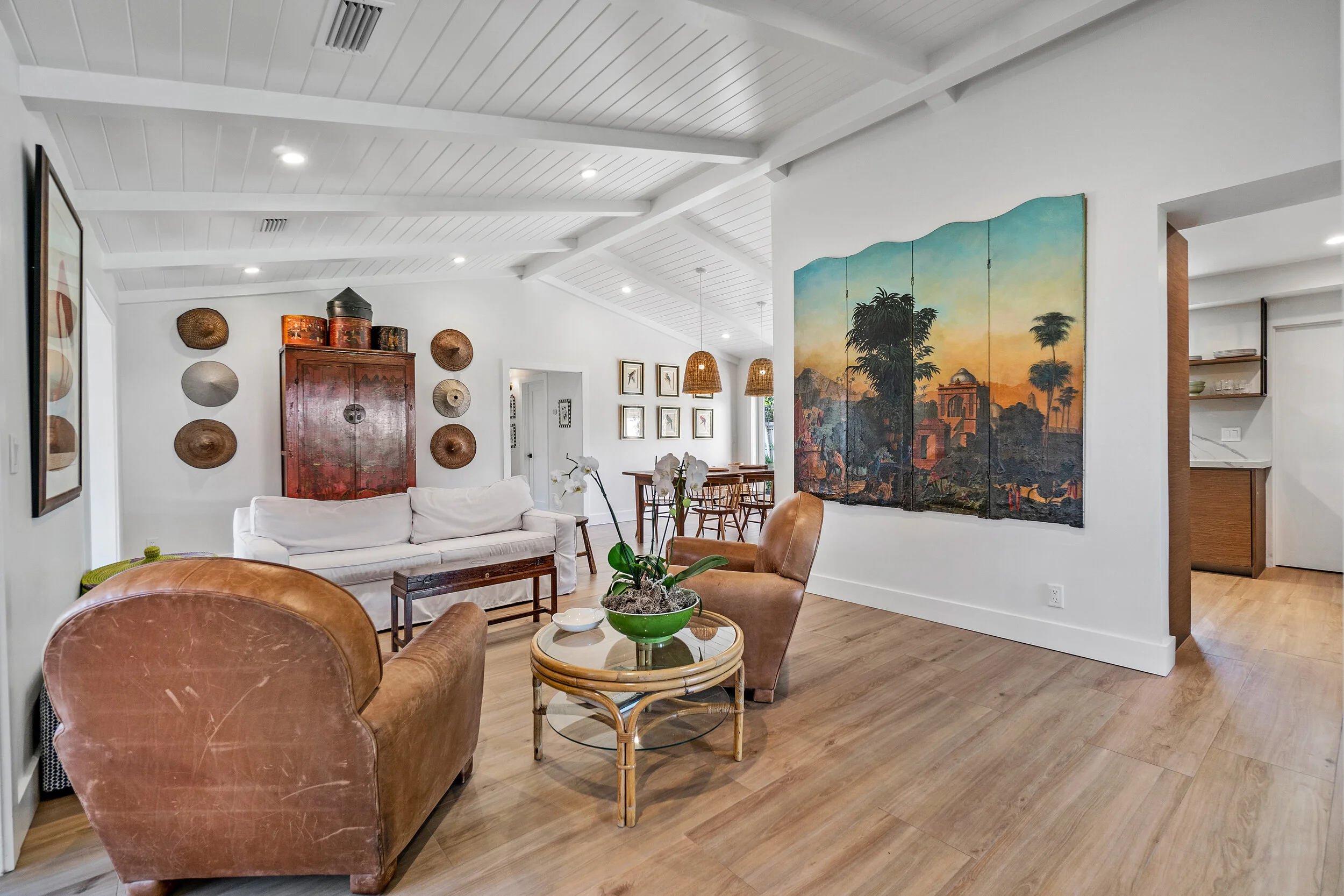 Renovated living room with white vaulted ceiling, leather chairs, white sofa, wood flooring, large wall art, and an open view toward the dining area.