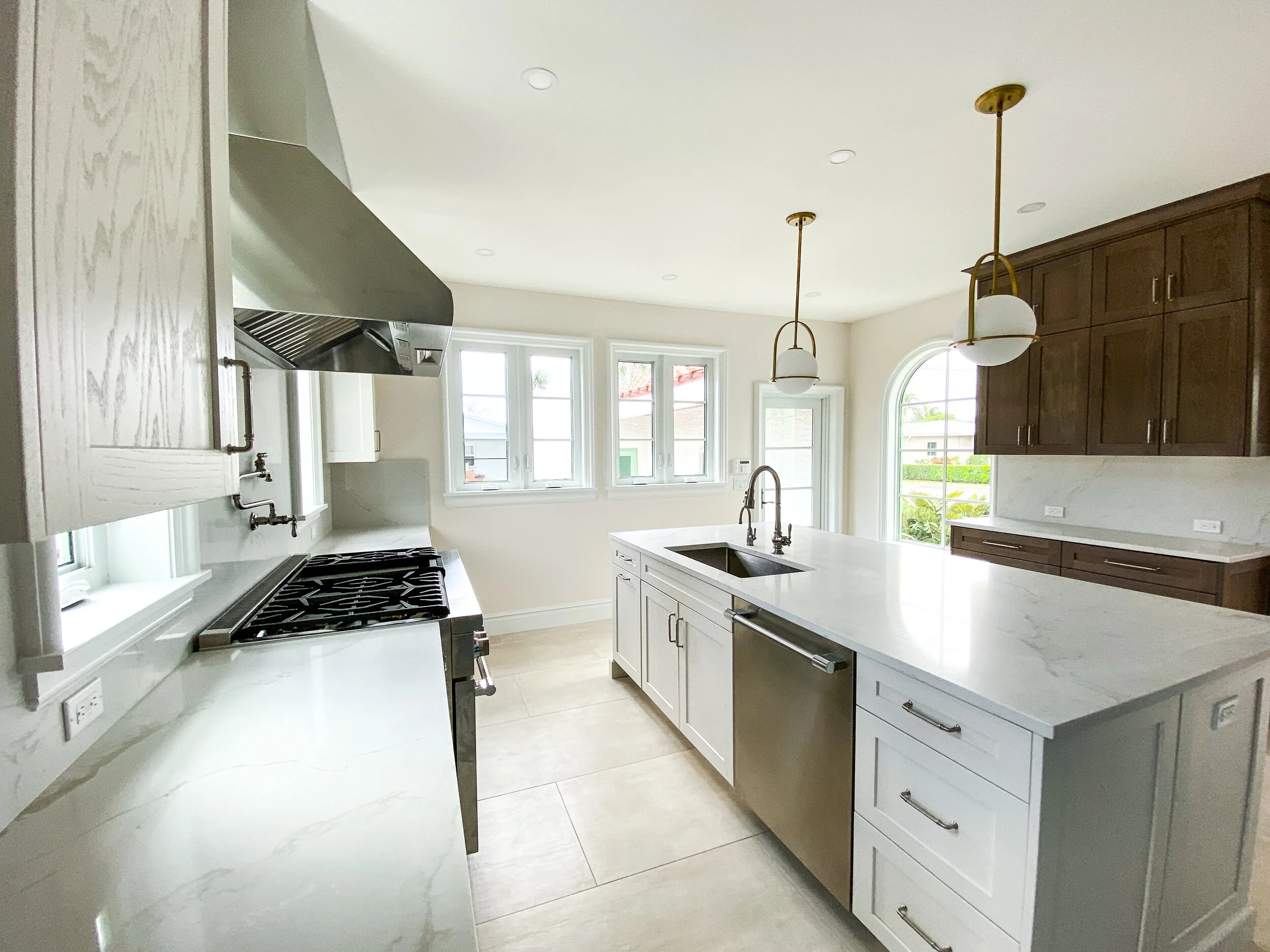 Renovated kitchen with white marble countertops, large island sink, stainless steel appliances, mixed white and wood cabinetry, arched window, and brass pendant lights.
