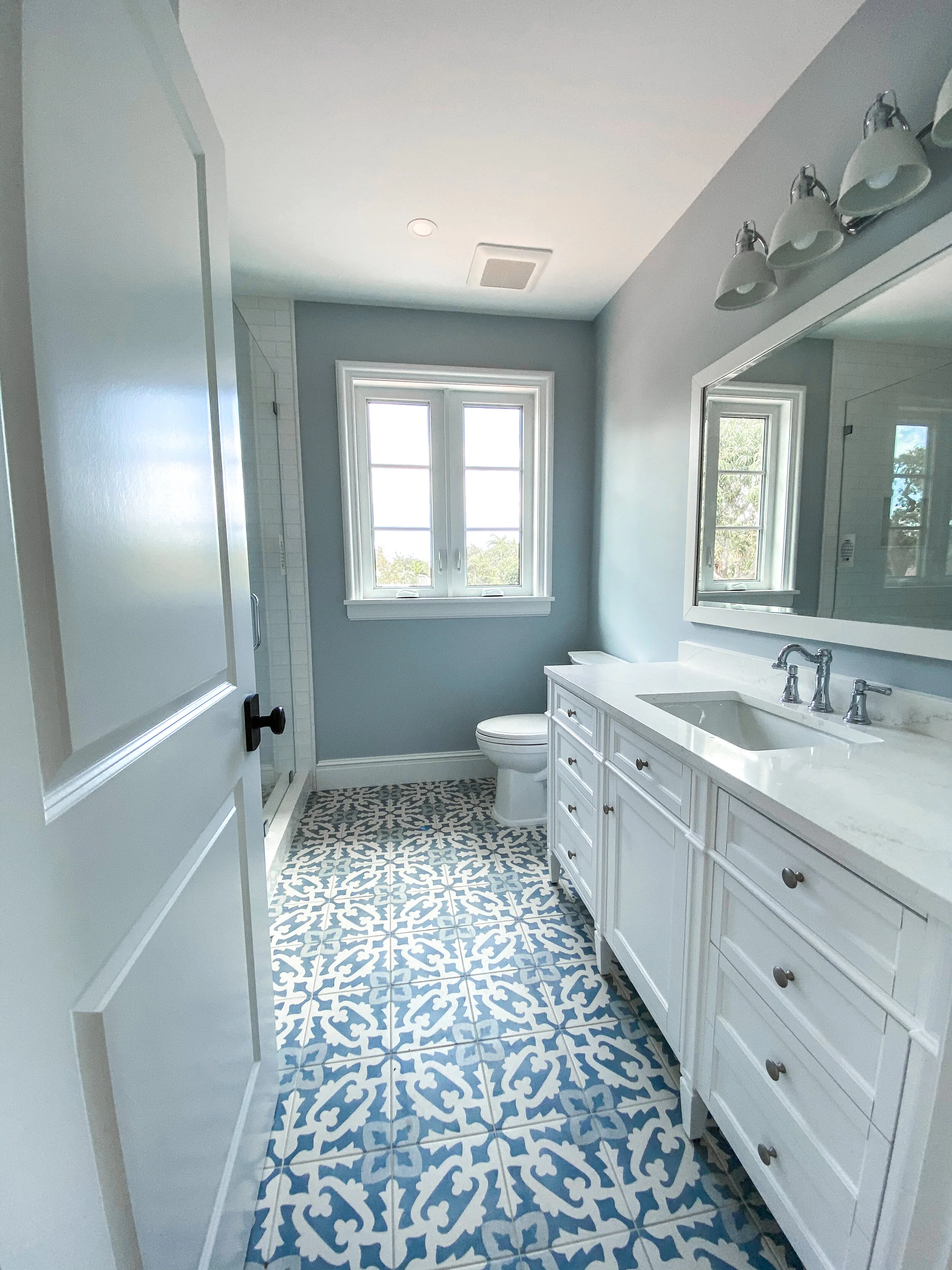 Bright bathroom with blue patterned tile floor, white double vanity, large wall mirror, walk-in shower, and two windows bringing in natural light.