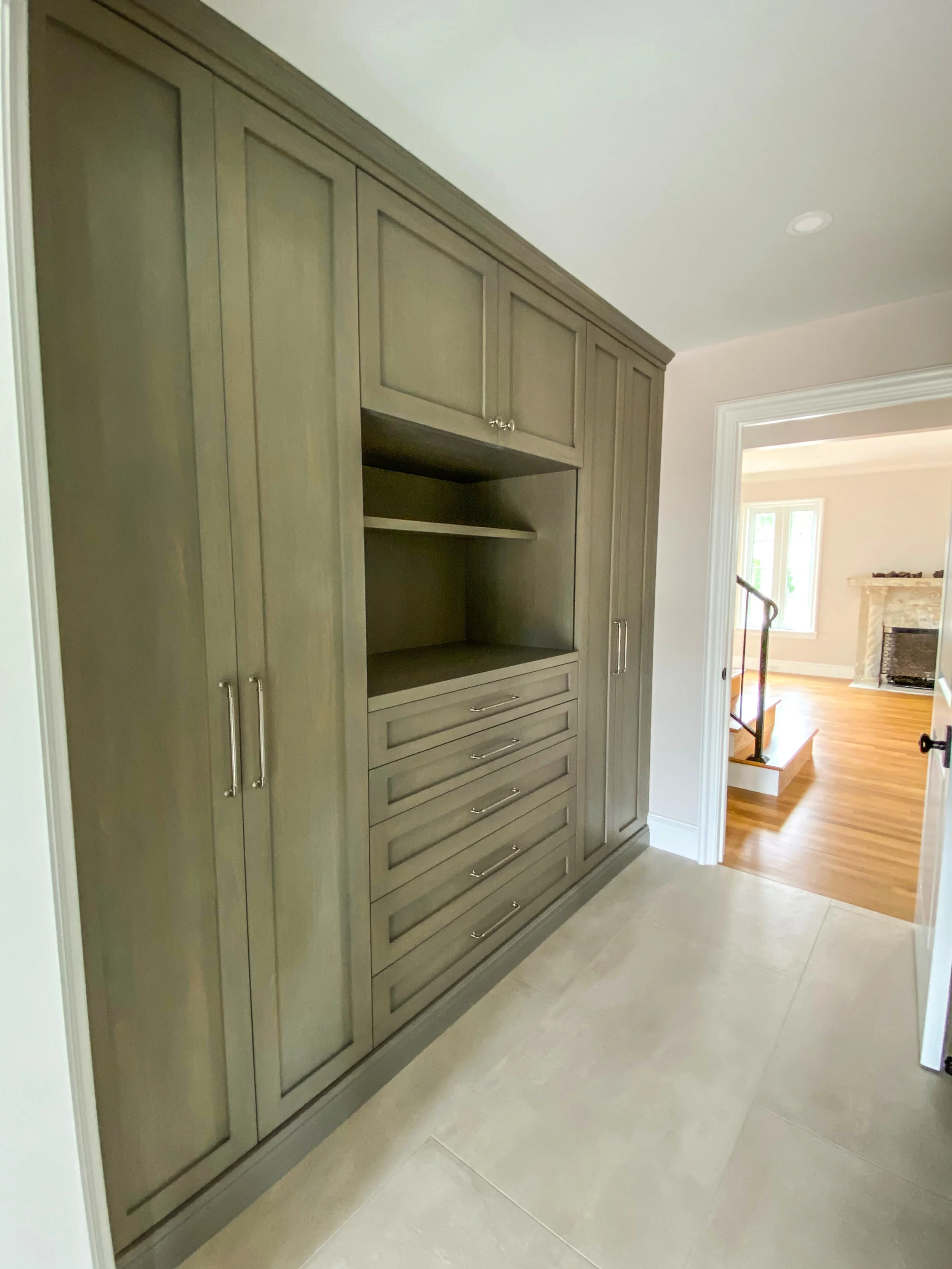 Custom built-in storage cabinet with tall paneled doors, drawers, open shelving, and a view into a bright room with hardwood floors.