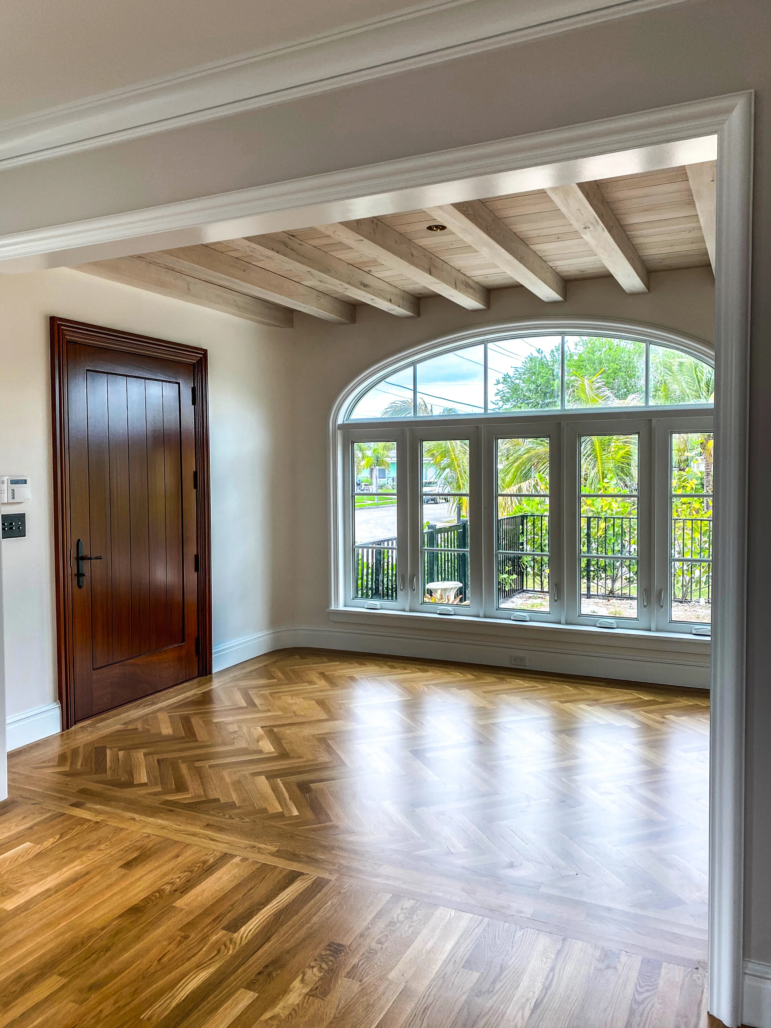 Historic-style interior room with herringbone wood floors, exposed ceiling beams, arched window wall, wood door, and natural light.