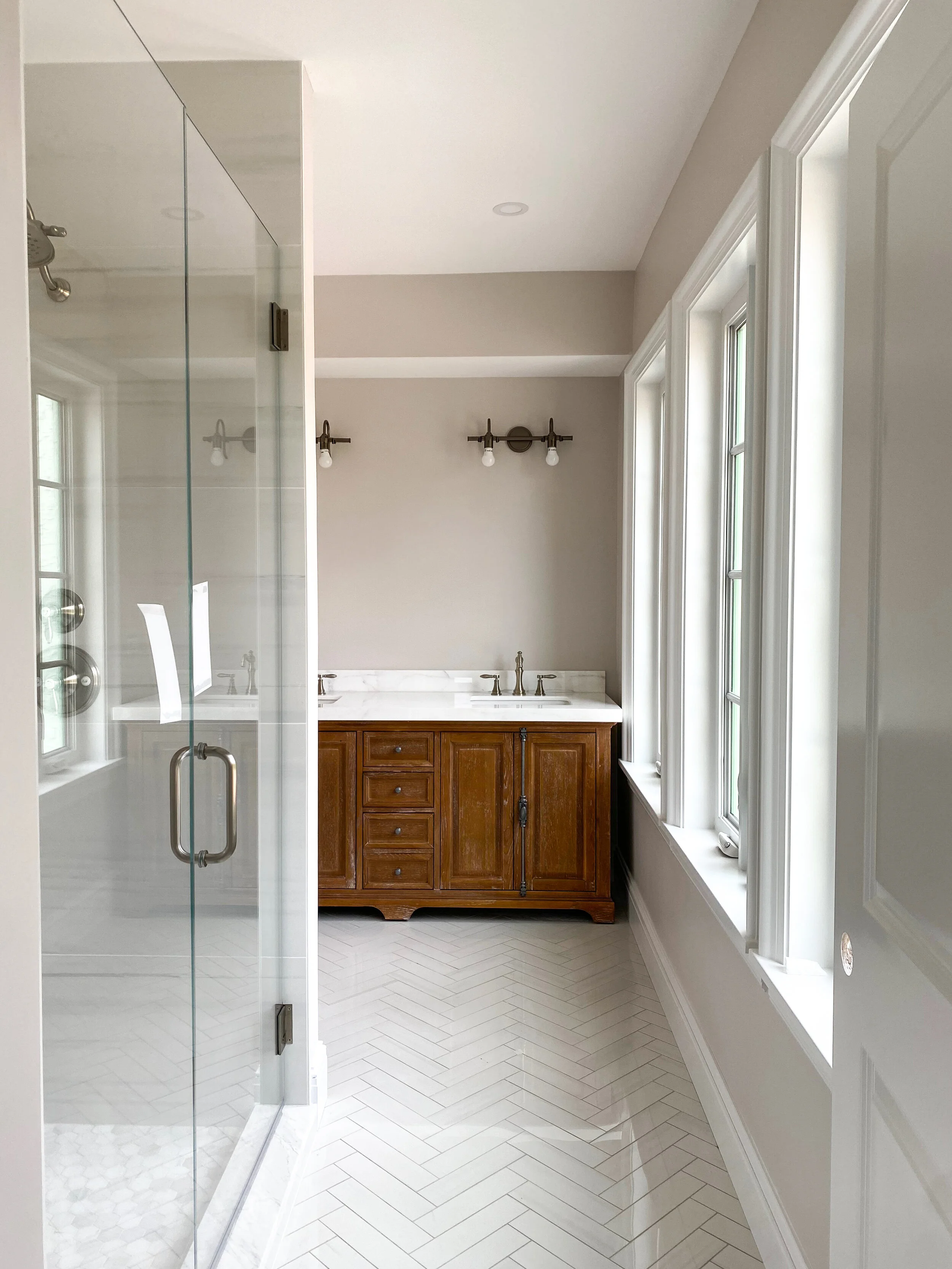 Bathroom with glass shower enclosure, white herringbone tile floor, wood vanity with marble countertop, wall sconces, and tall windows.