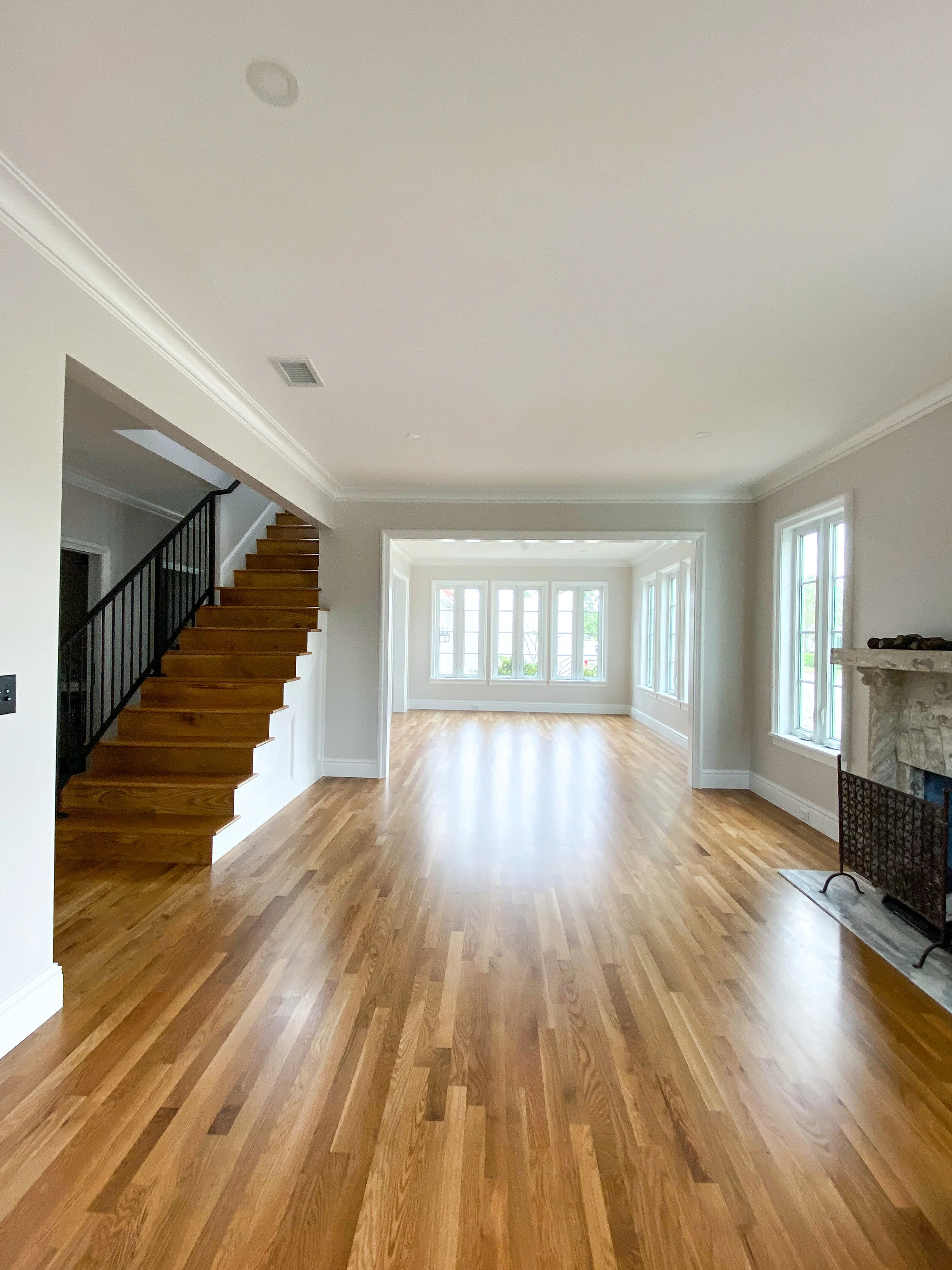 Open living room with refinished hardwood floors, staircase with black railing, fireplace, large windows, and bright neutral walls.