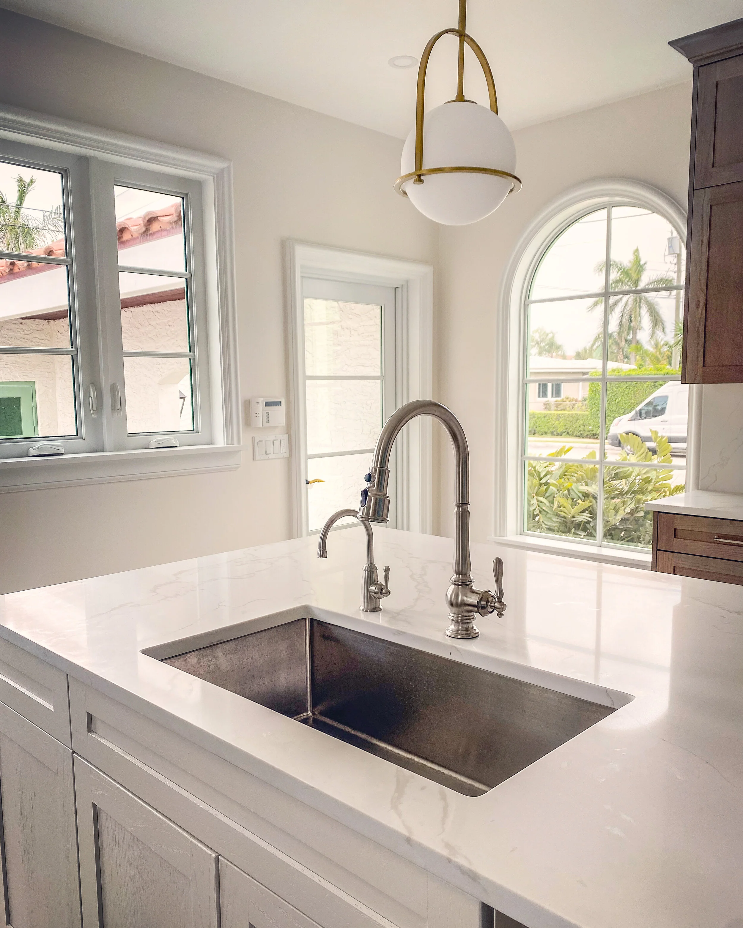 Close-up of renovated kitchen island with marble countertop, undermount sink, polished faucet, brass pendant light, arched window, and wood cabinetry.