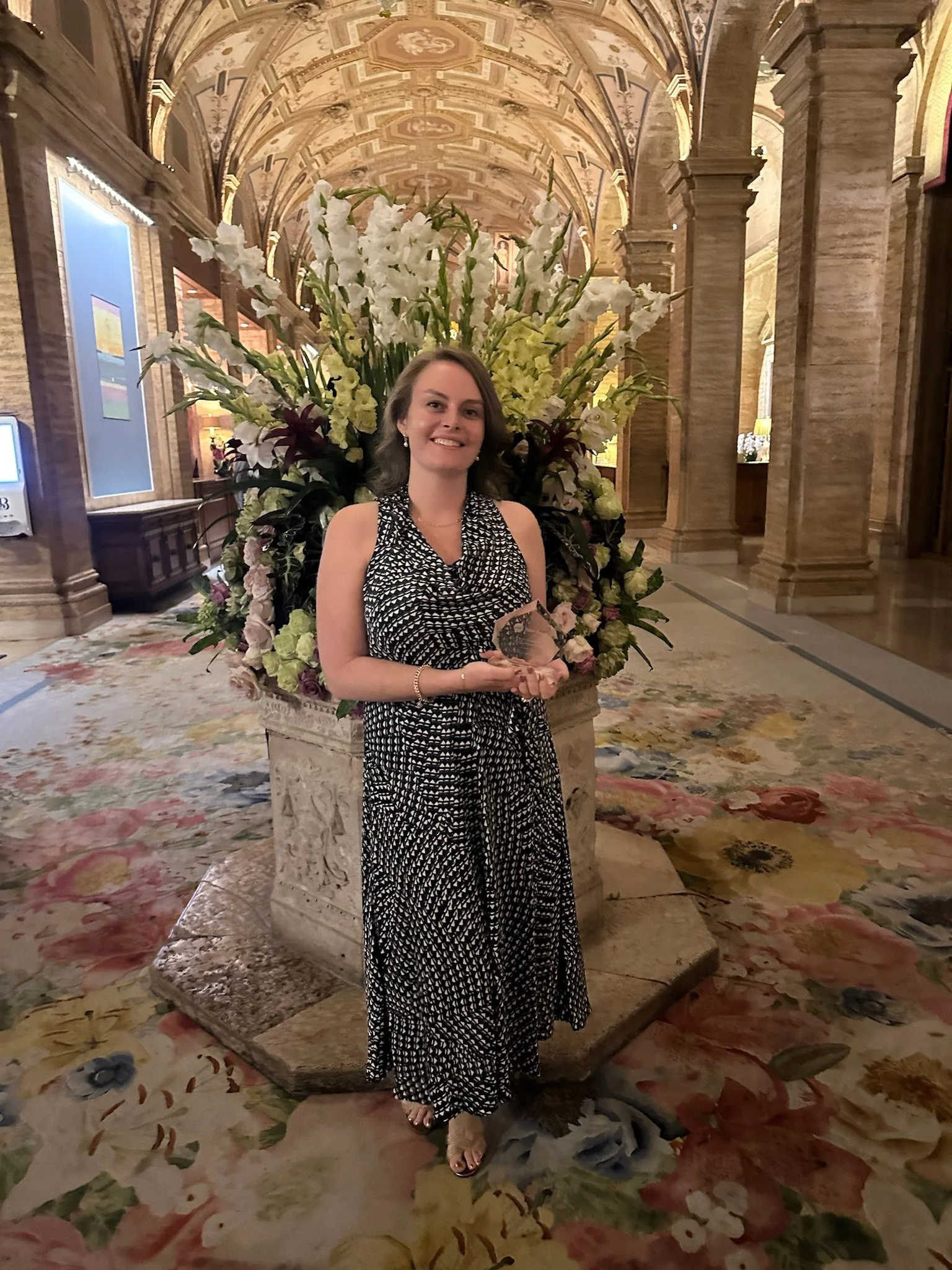 A woman standing indoors in front of a large floral arrangement, smiling and holding an award or trophy, in a grand, ornately decorated hall with high arched ceilings and intricate architectural details.