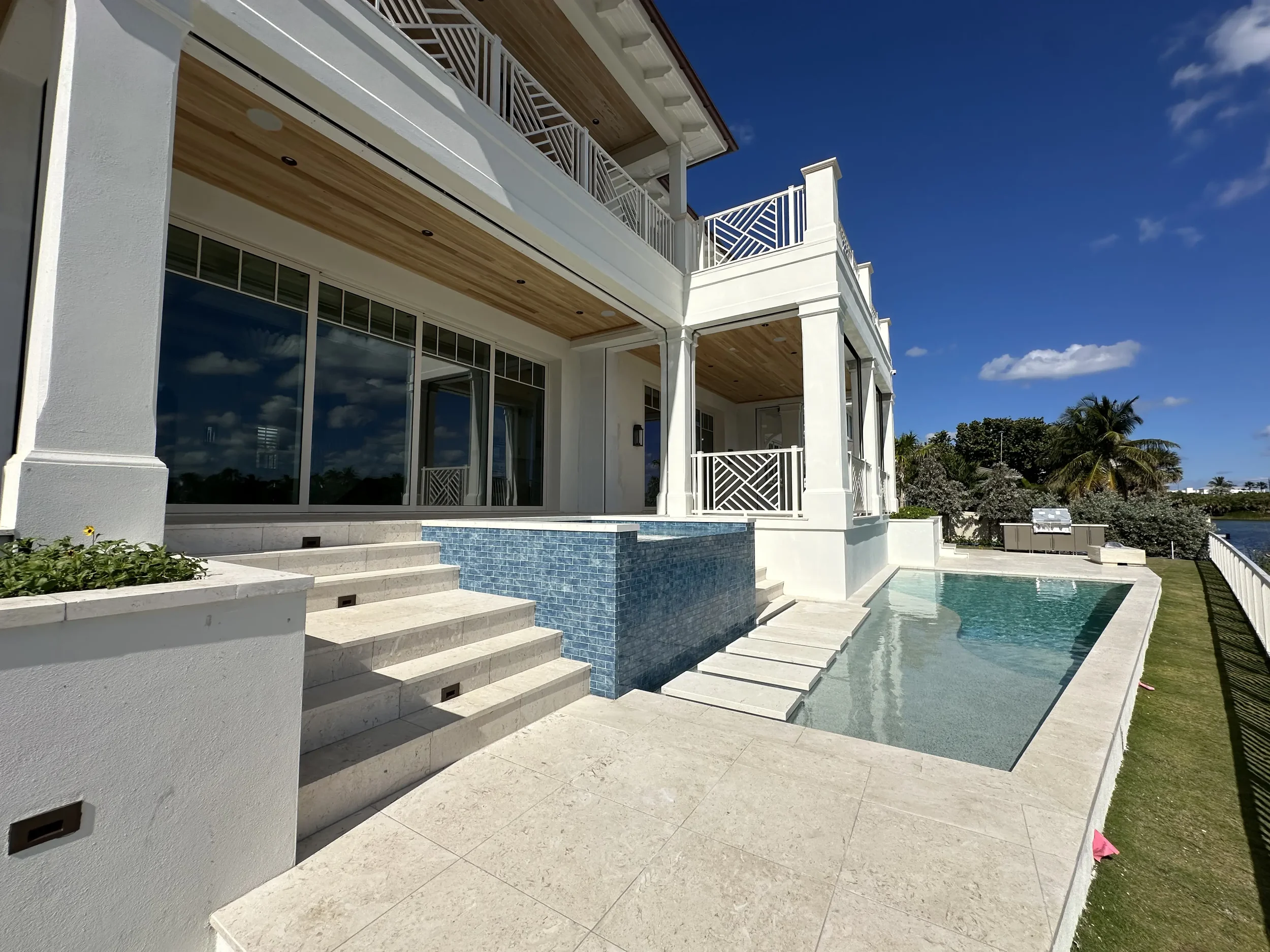 Outdoor pool and spa area beside a white waterfront home with glass doors, covered balcony, blue tile feature wall, and stone pavers.