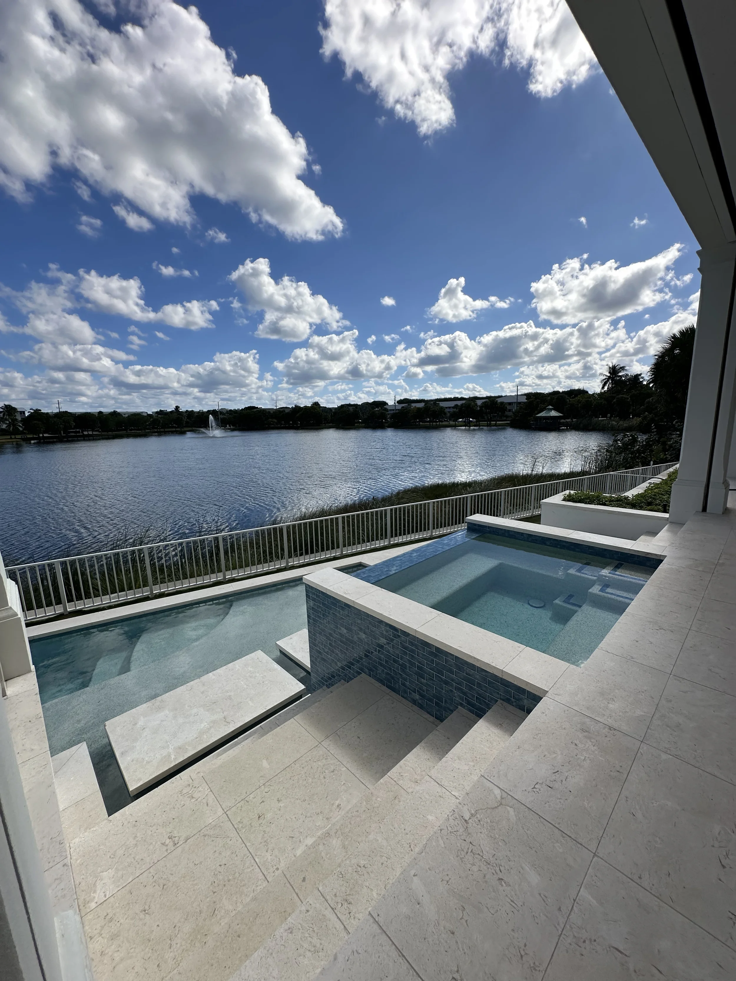 View from covered patio over a pool and raised spa toward a lake with a fountain, white railing, and cloudy blue sky.