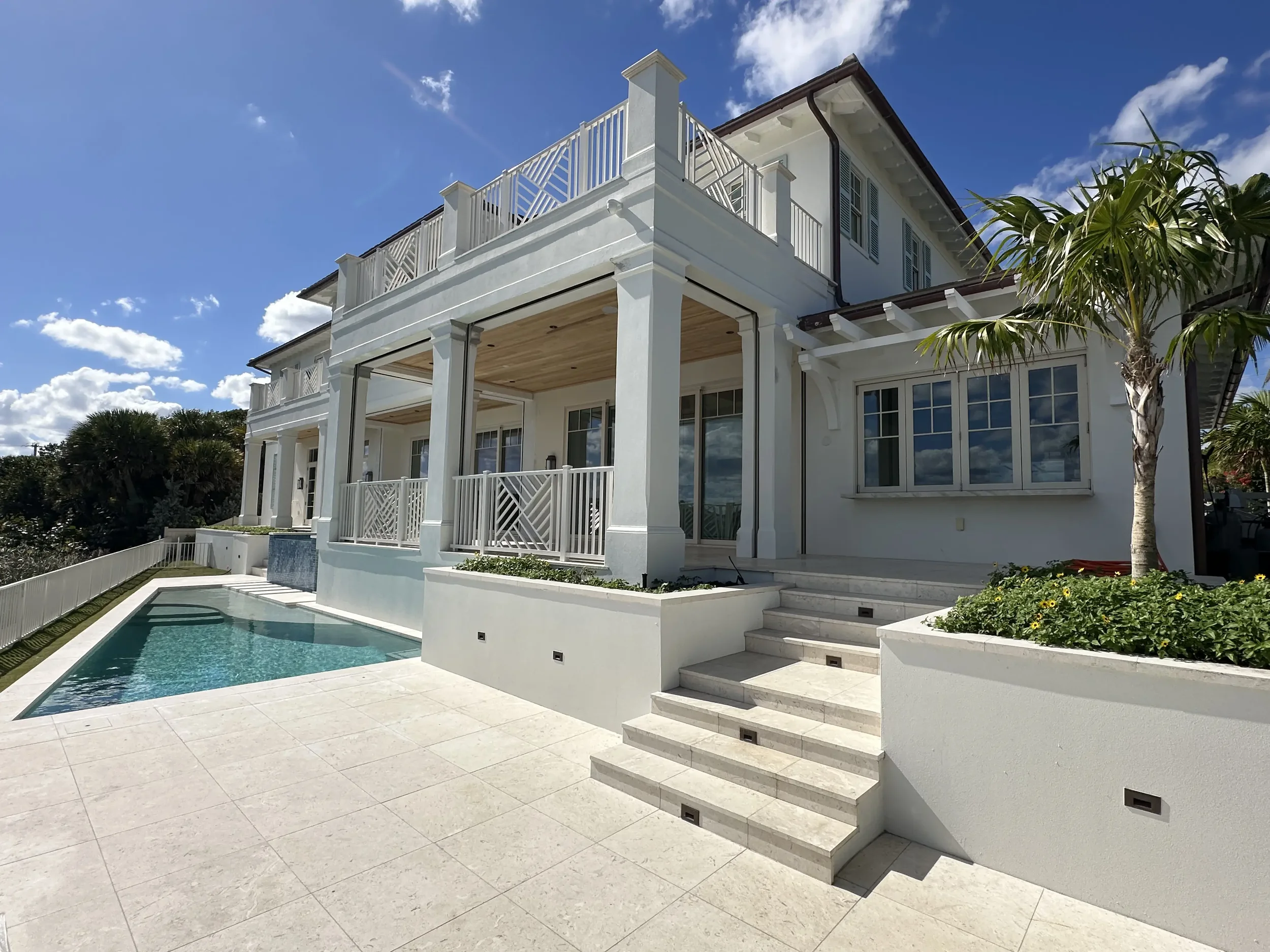 Rear exterior of a large white waterfront home with covered terrace, upper balcony, pool, stone steps, and palm tree landscaping.