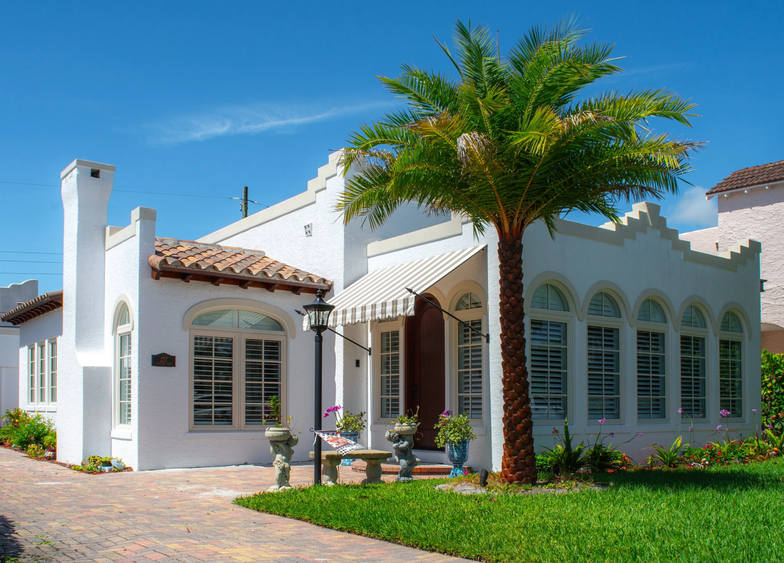 A white house with large windows, a tiled roof, and a front yard with a palm tree, colorful flowers, and outdoor furniture, under a clear blue sky.
