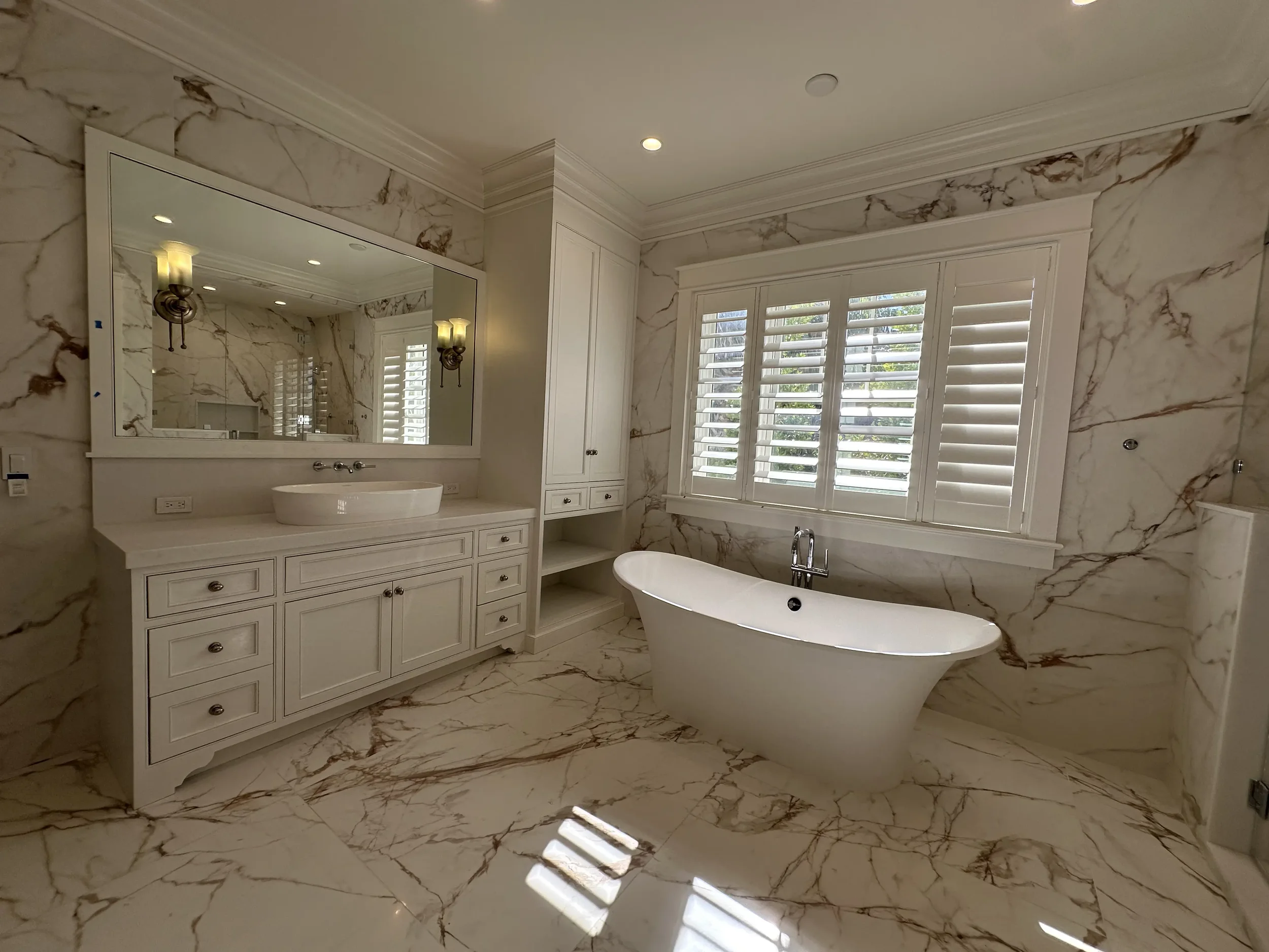 Bright marble bathroom with freestanding bathtub, double vanity, large mirror, wall sconces, shutters, and marble-look floor and wall surfaces.