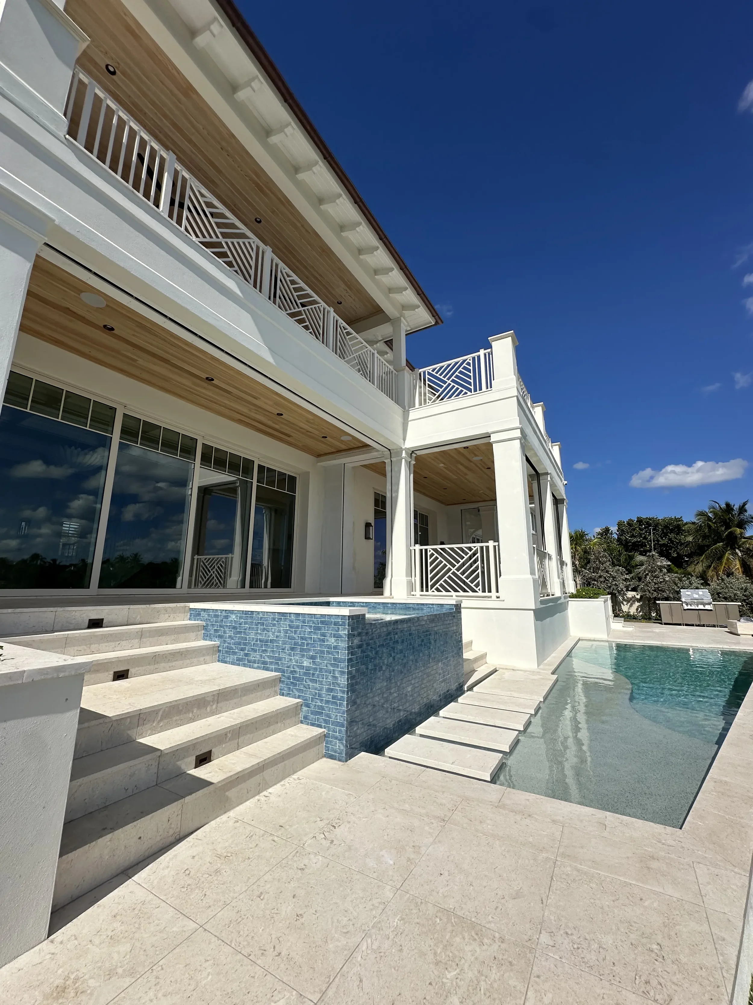 Low-angle view of a white waterfront home with covered balconies, decorative railings, blue tile spa wall, and poolside stone steps.