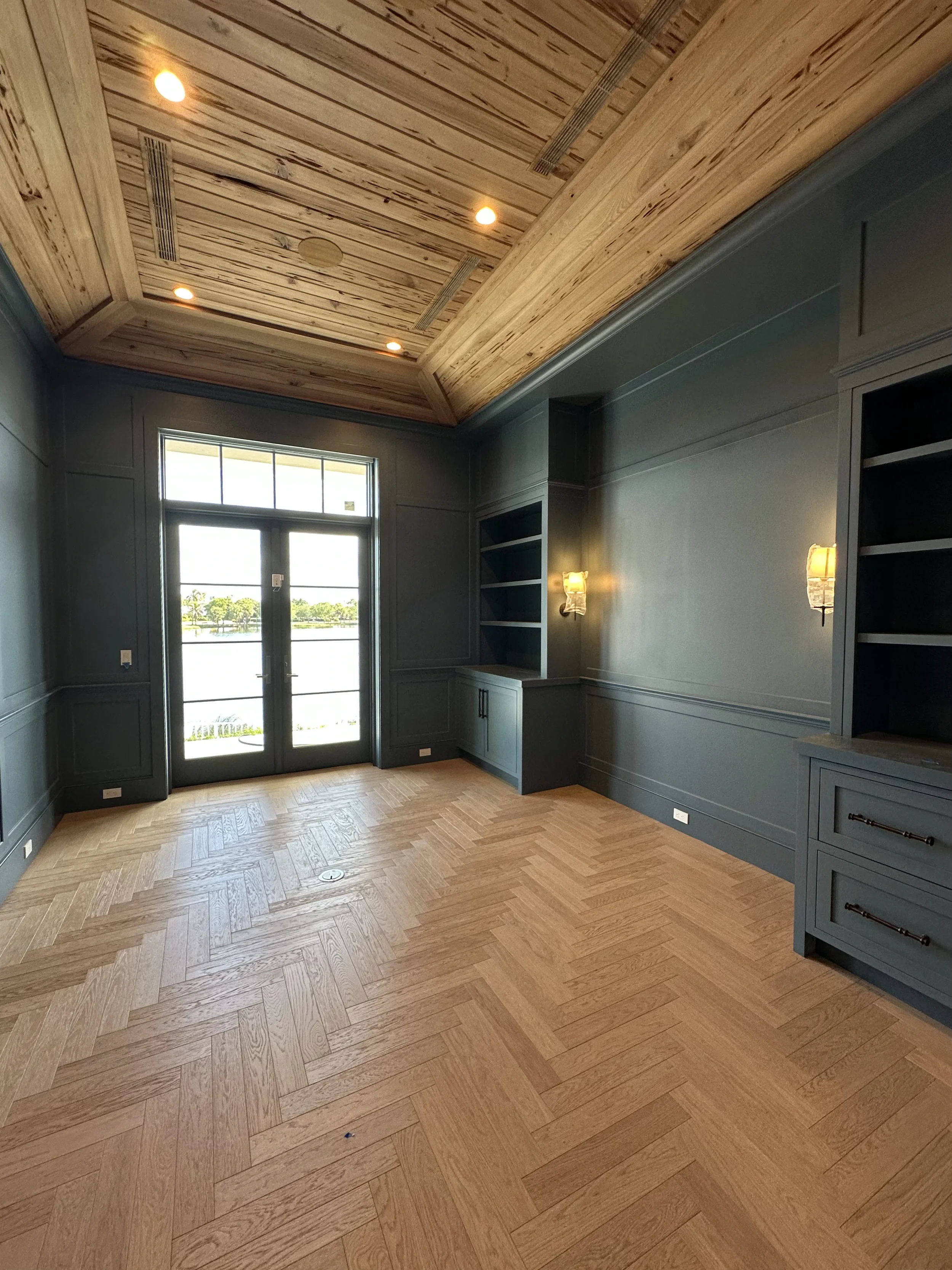 Dark paneled study with herringbone wood flooring, built-in shelving, wall sconces, French doors, and a detailed wood ceiling.