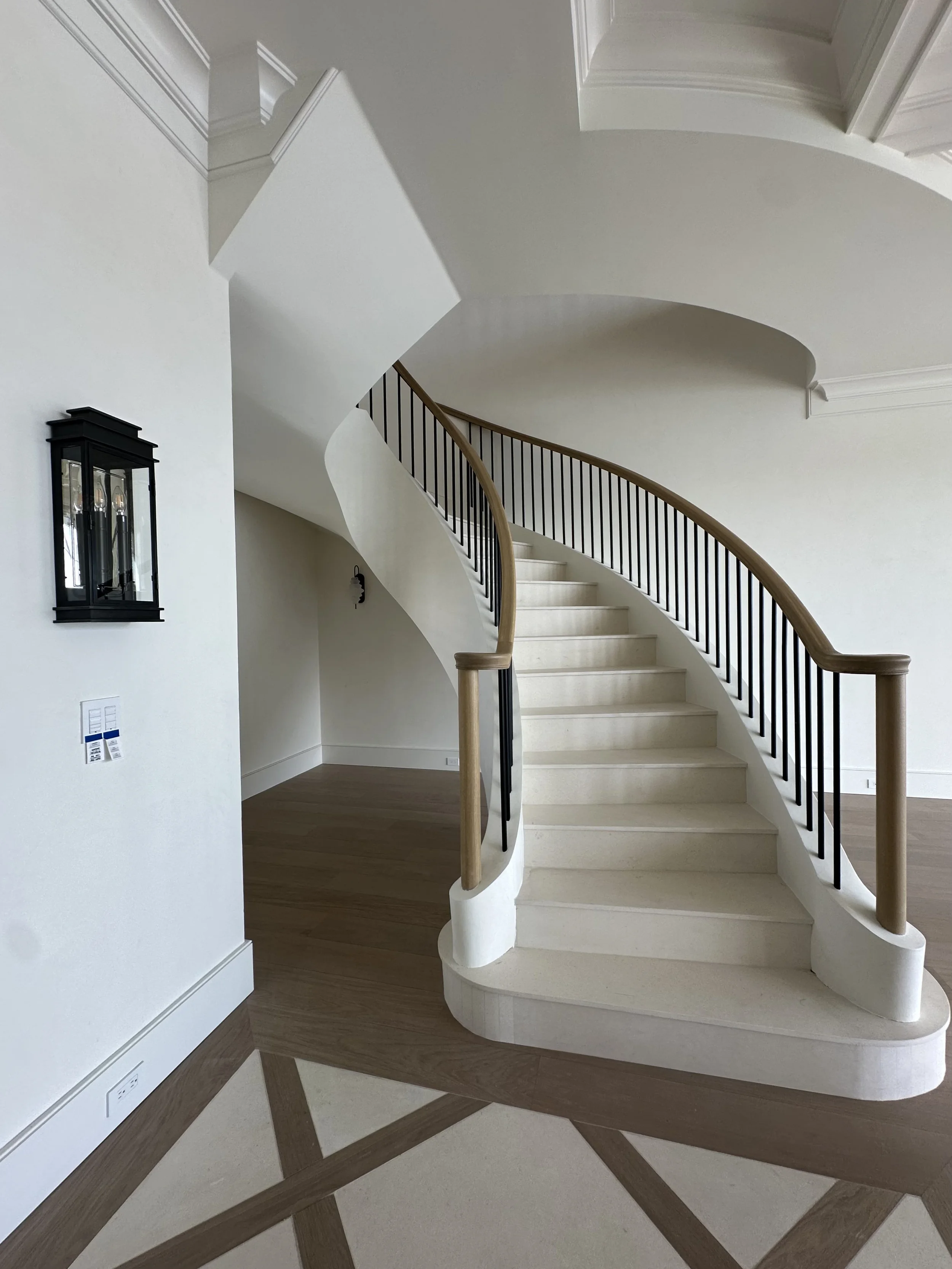 Grand foyer with curved staircase, arched wood entry doors, black stair railings, wood handrail, and patterned floor inlay.