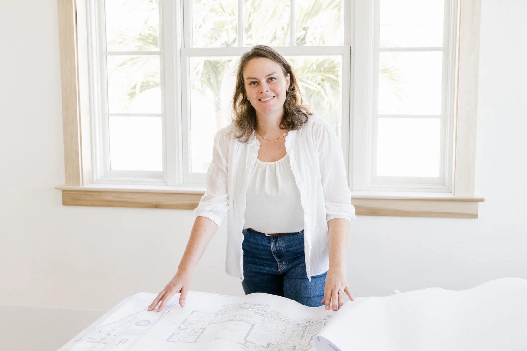 A woman with brown hair, wearing a white blouse and blue jeans, standing at a table with architectural plans, in front of a large window with white framing, bright natural light coming in.