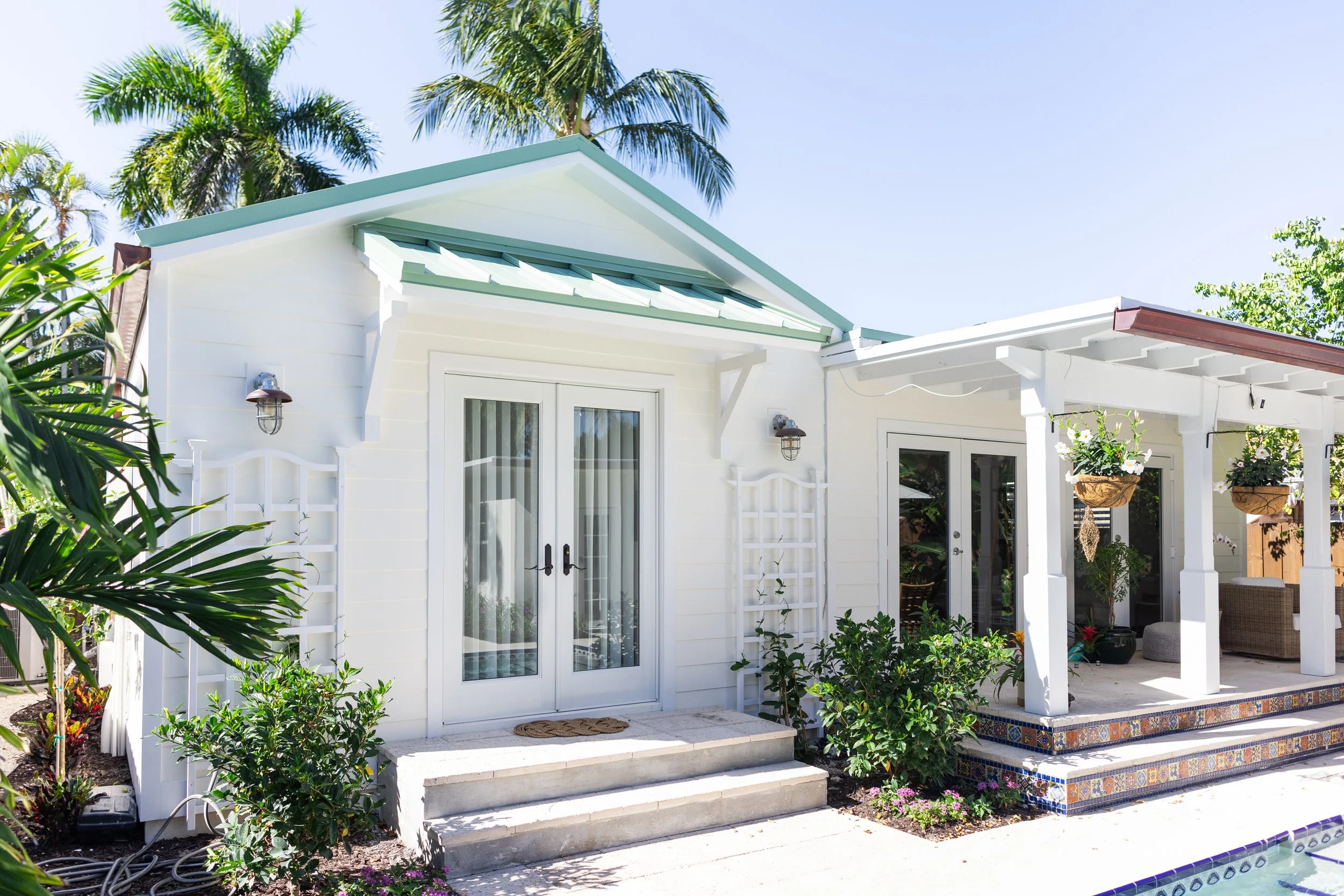A white house with a glass door, surrounded by green plants, palm trees, and a concrete staircase leading to a covered patio area with hanging plants and outdoor furniture. The house has a green metal roof and white wooden siding.