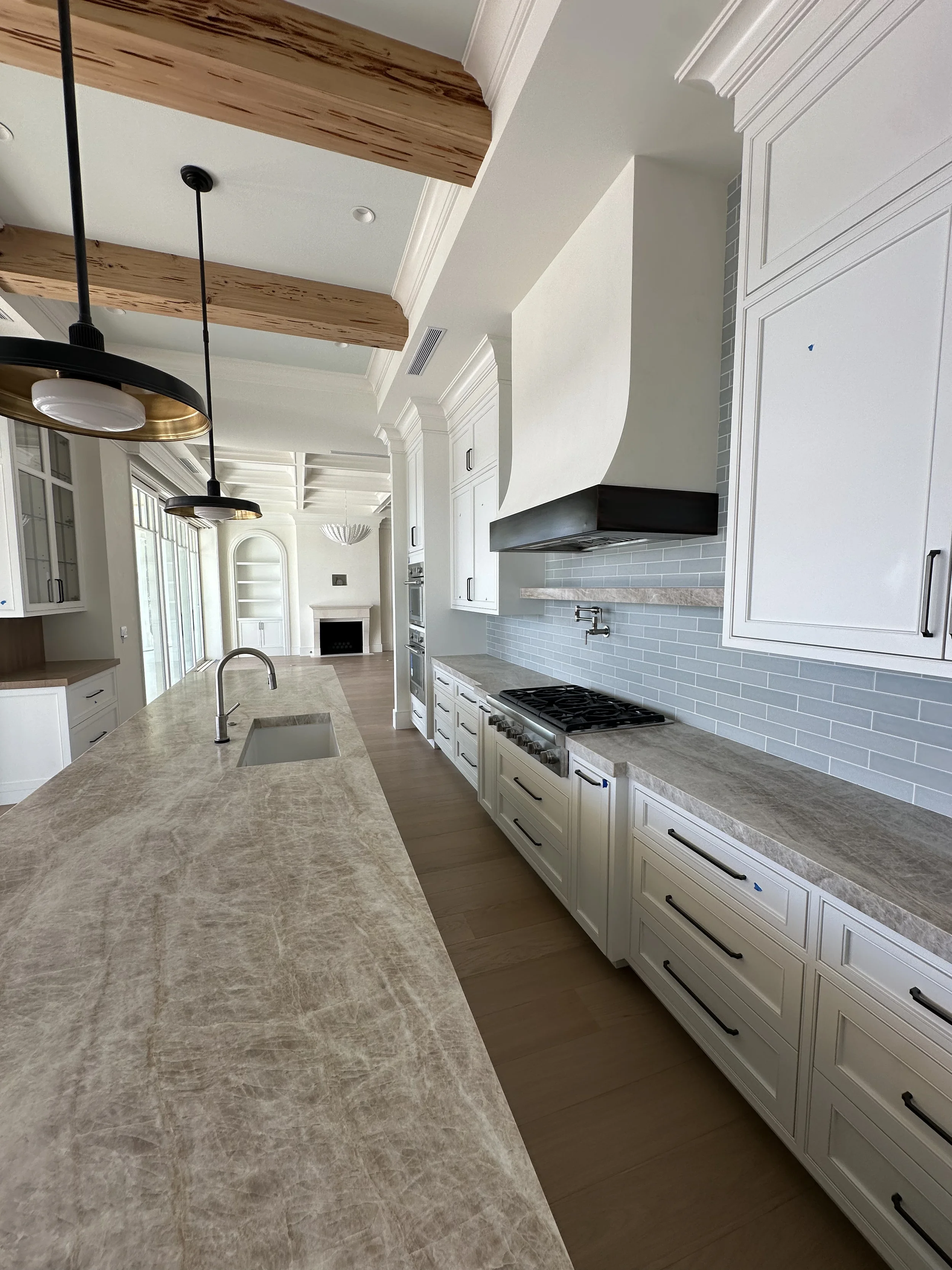 Waterfront kitchen with long stone island, exposed wood beams, black pendant lights, white cabinetry, gas range, and fireplace beyond.