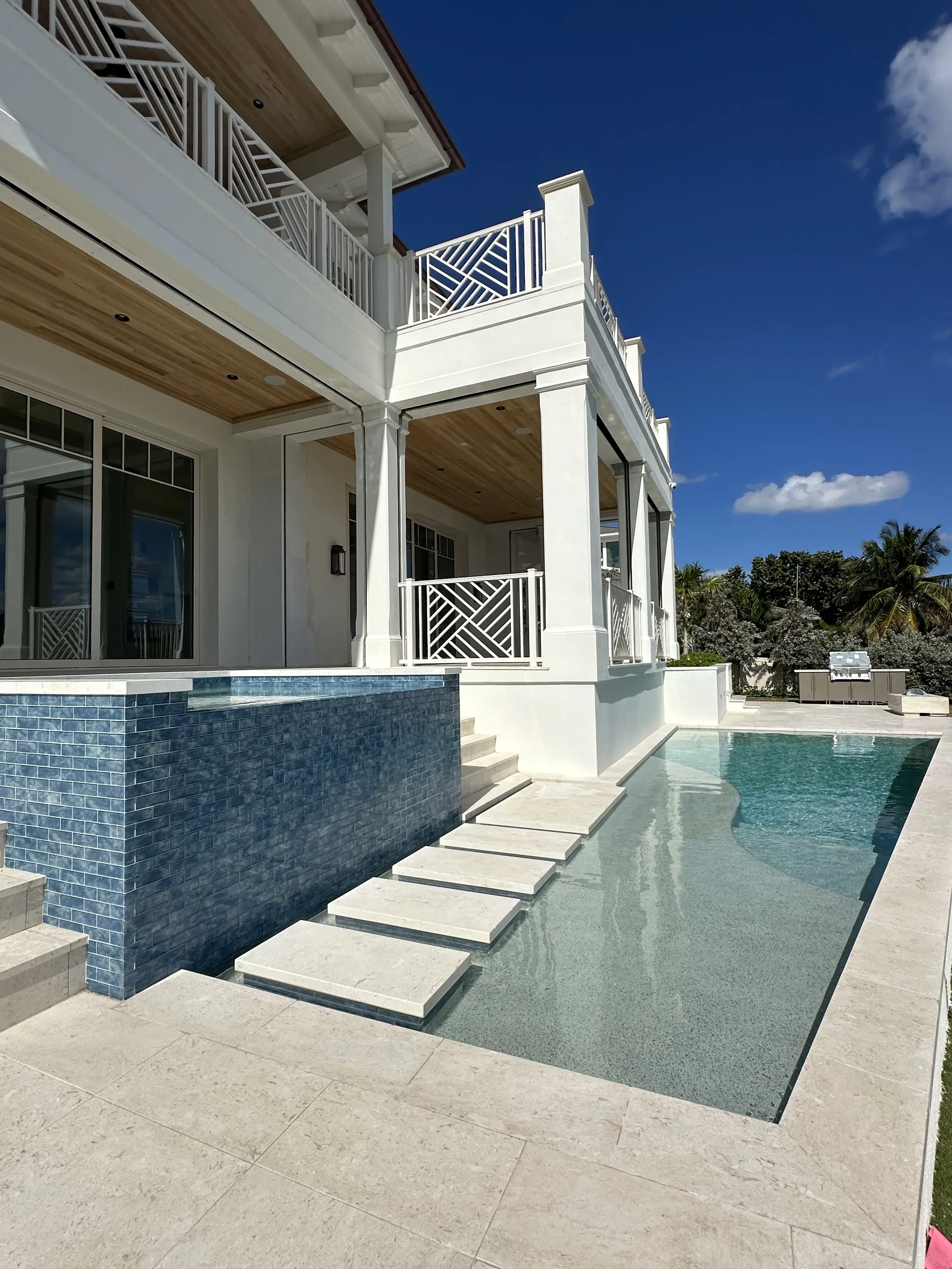 Waterfront pool area with blue tile spa wall, stepping stones, white covered balconies, decorative railings, and bright stone decking.