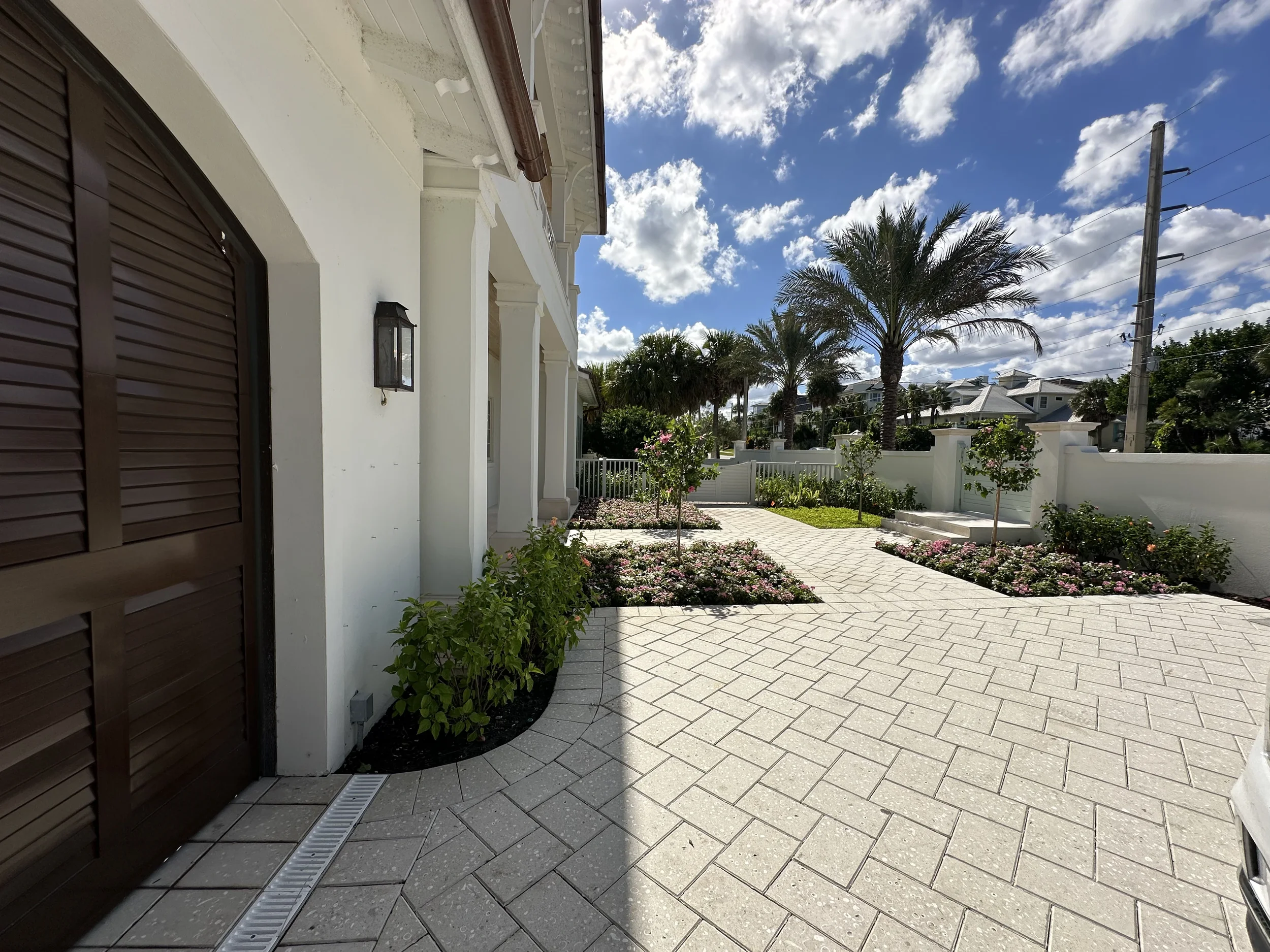 Front walkway of a white coastal home with arched wood door, paved courtyard, landscaped beds, palm tree, and sunny blue sky.