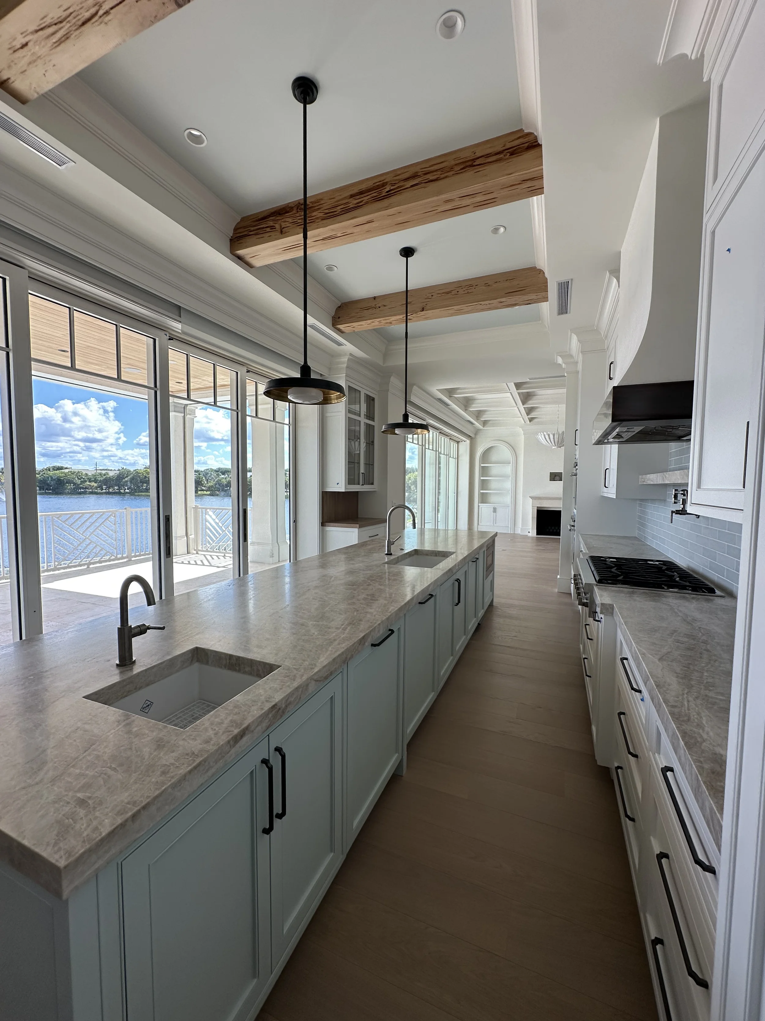 Long waterfront kitchen with large stone island, dual sinks, black pendant lights, exposed wood beams, and glass doors overlooking the water.