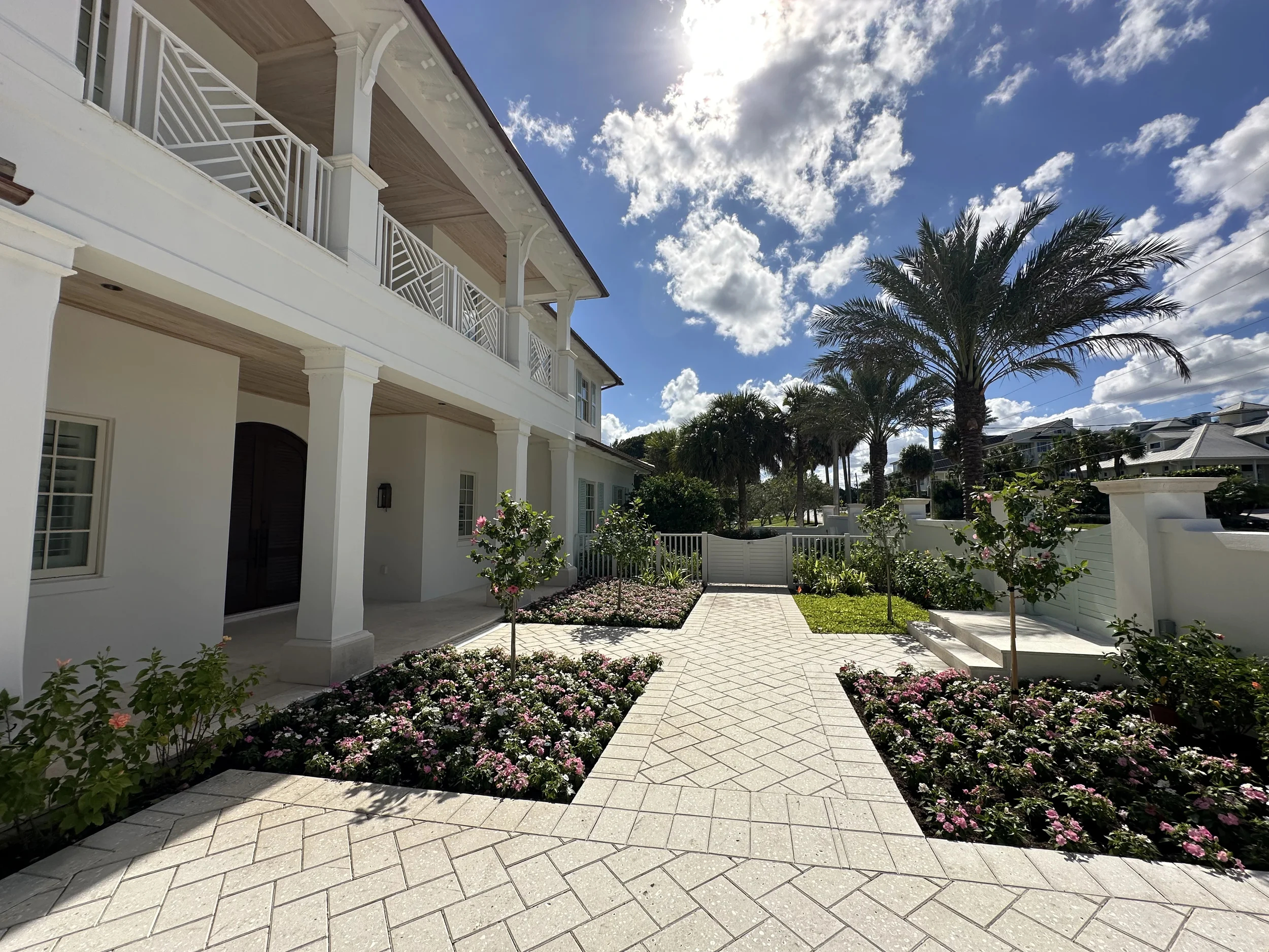 Front courtyard of a white coastal home with paved walkway, flower beds, palm trees, covered entry, and upper balcony.