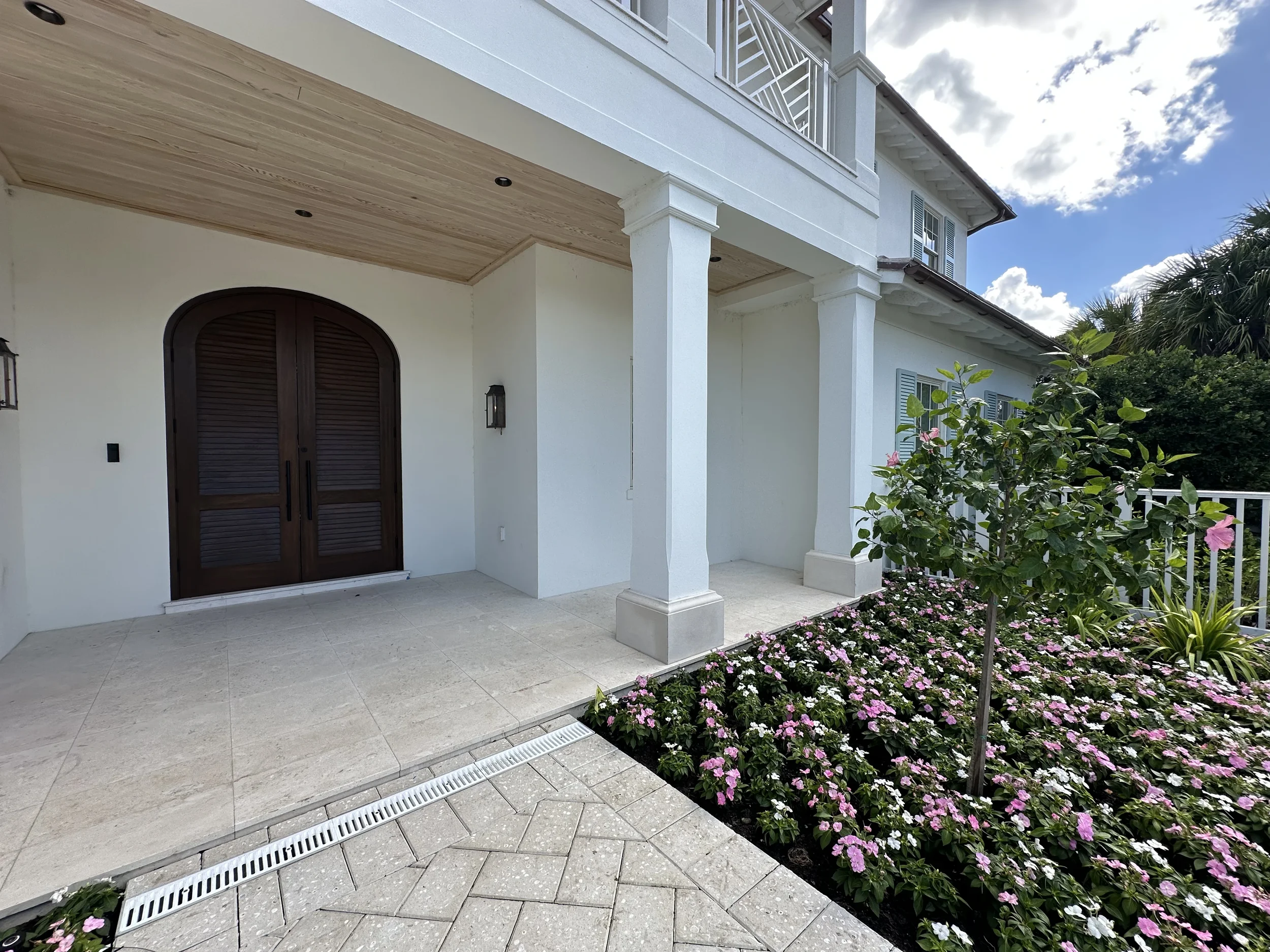 Covered front entry of a waterfront home with arched wood double doors, white columns, stone pavers, and landscaped flower beds.