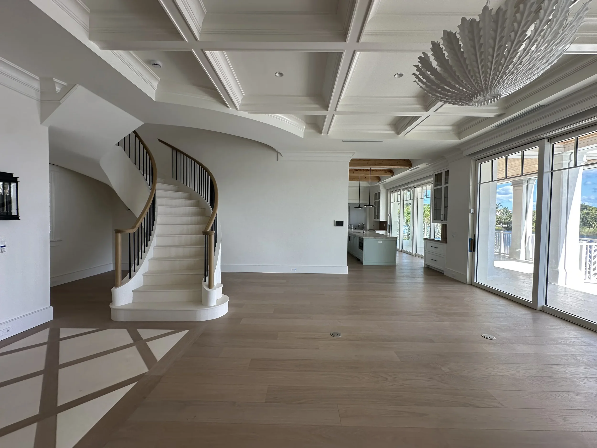 Open living area with curved staircase, coffered ceiling, large chandelier, wood flooring, glass doors, and view toward the kitchen.