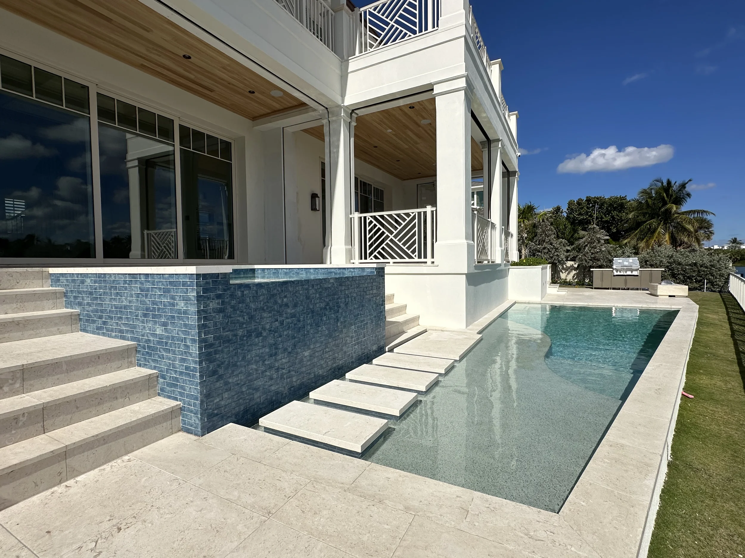 Rear exterior of a waterfront home with covered balconies, white railings, blue tile spa wall, swimming pool, and stone patio.