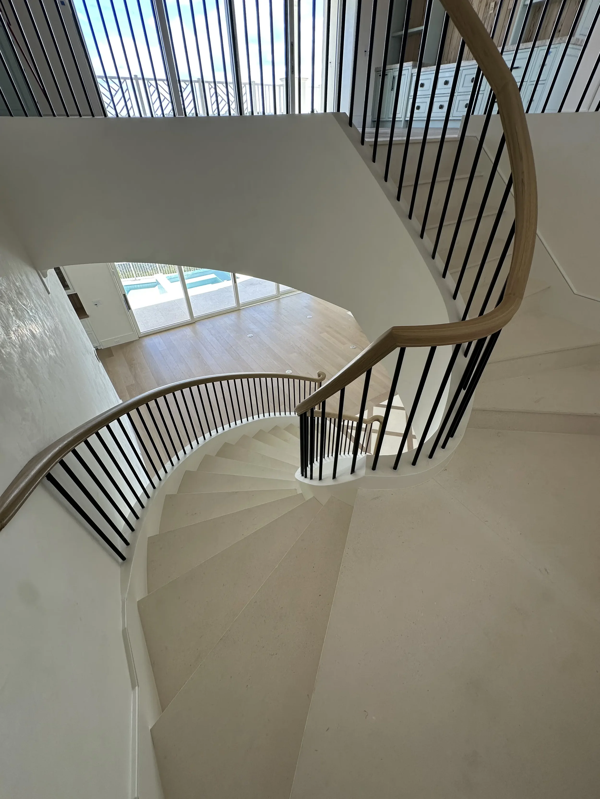 View down a curved staircase with black balusters and wood handrail toward a bright living area with glass doors and pool outside.