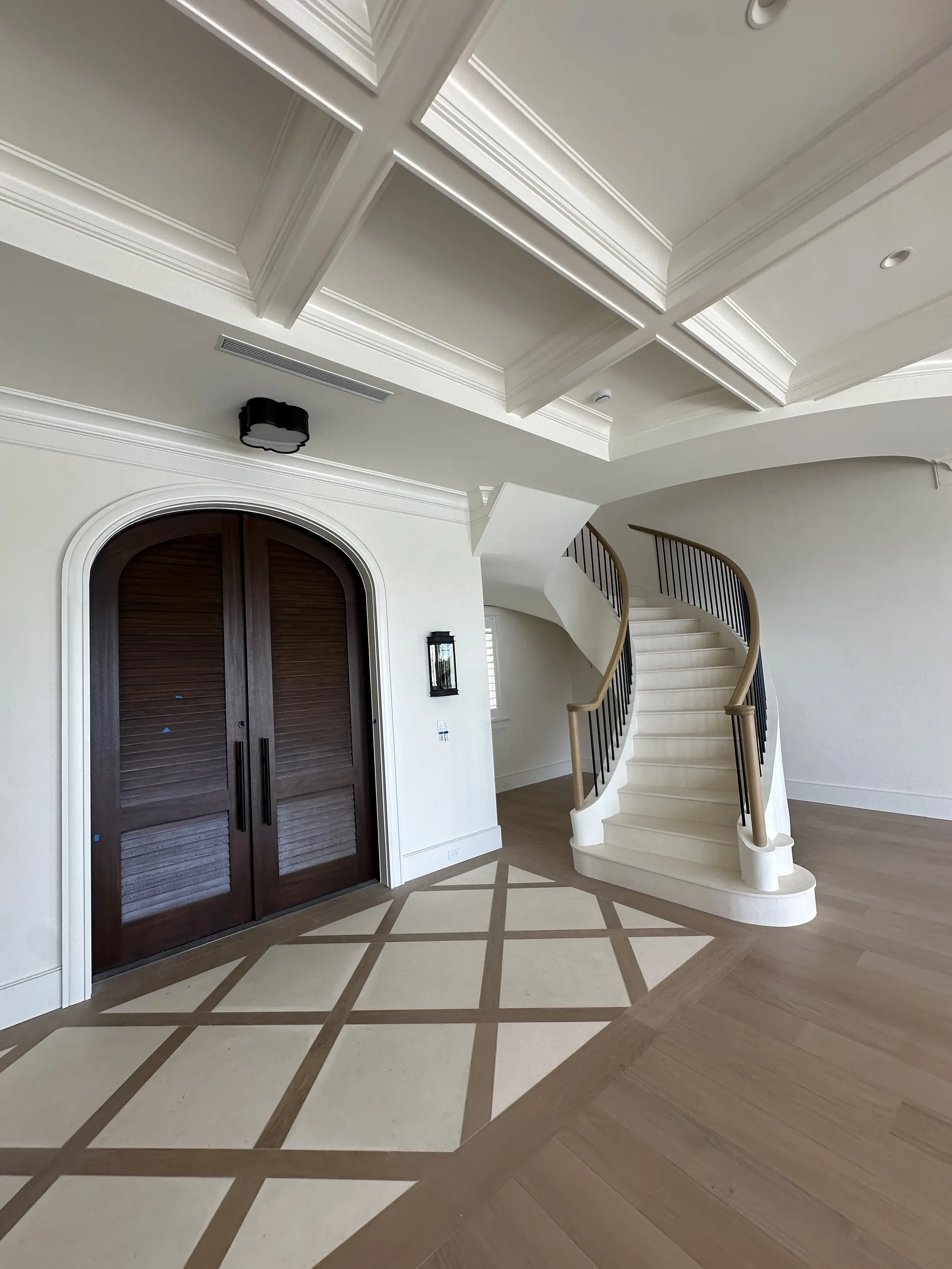 Elegant foyer with arched wood double doors, curved staircase, coffered ceiling, patterned floor inlay, and wall lanterns.