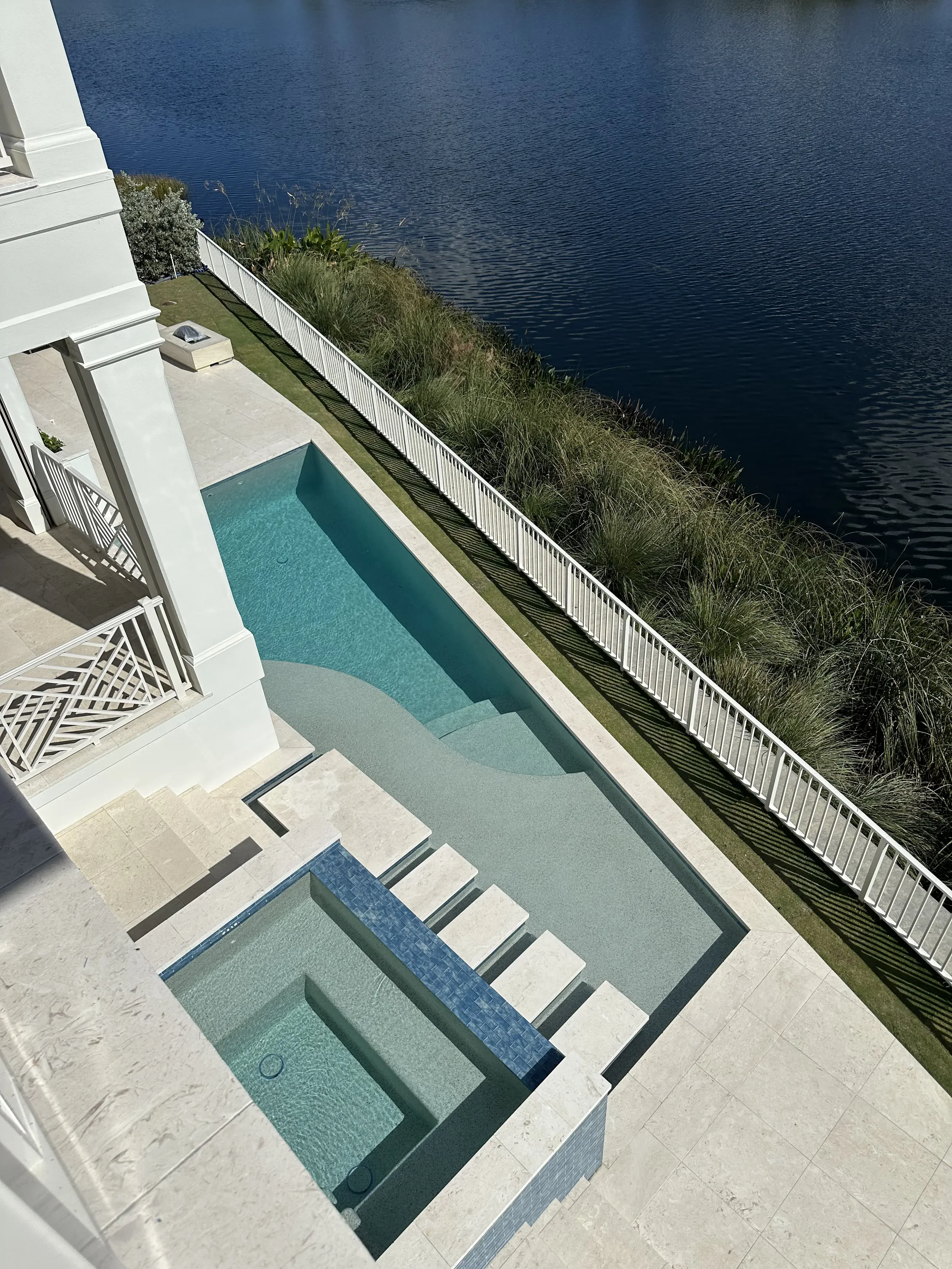 Overhead view of a waterfront pool and raised spa beside a white home, with stepping stones, blue tile wall, and lake beyond.