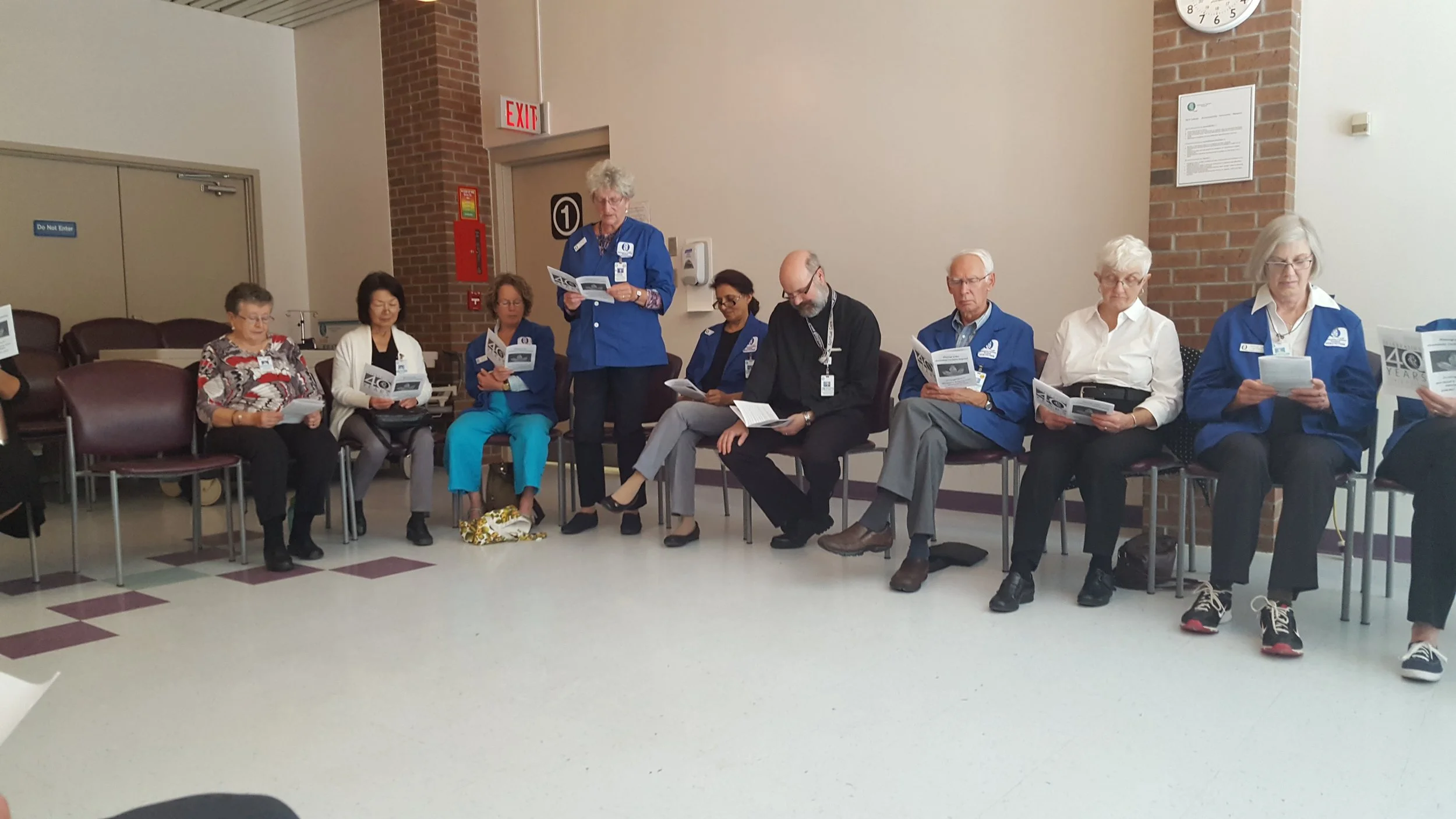 A group of nine people, are sitting in a semi-circle in be a meeting room. Several are wearing the volunteer blue vests and holding booklets, they are participating in a spiritual