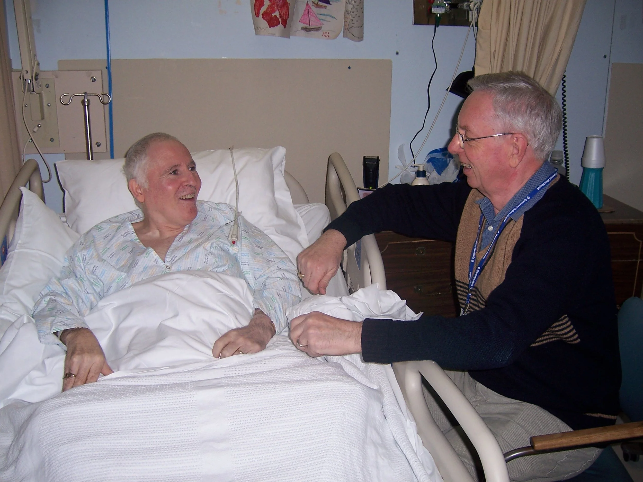 A man in a hospital bed, wearing a gown and smiling, is talking with a  chaplain who is sitting beside the bed and leaning in toward him.
