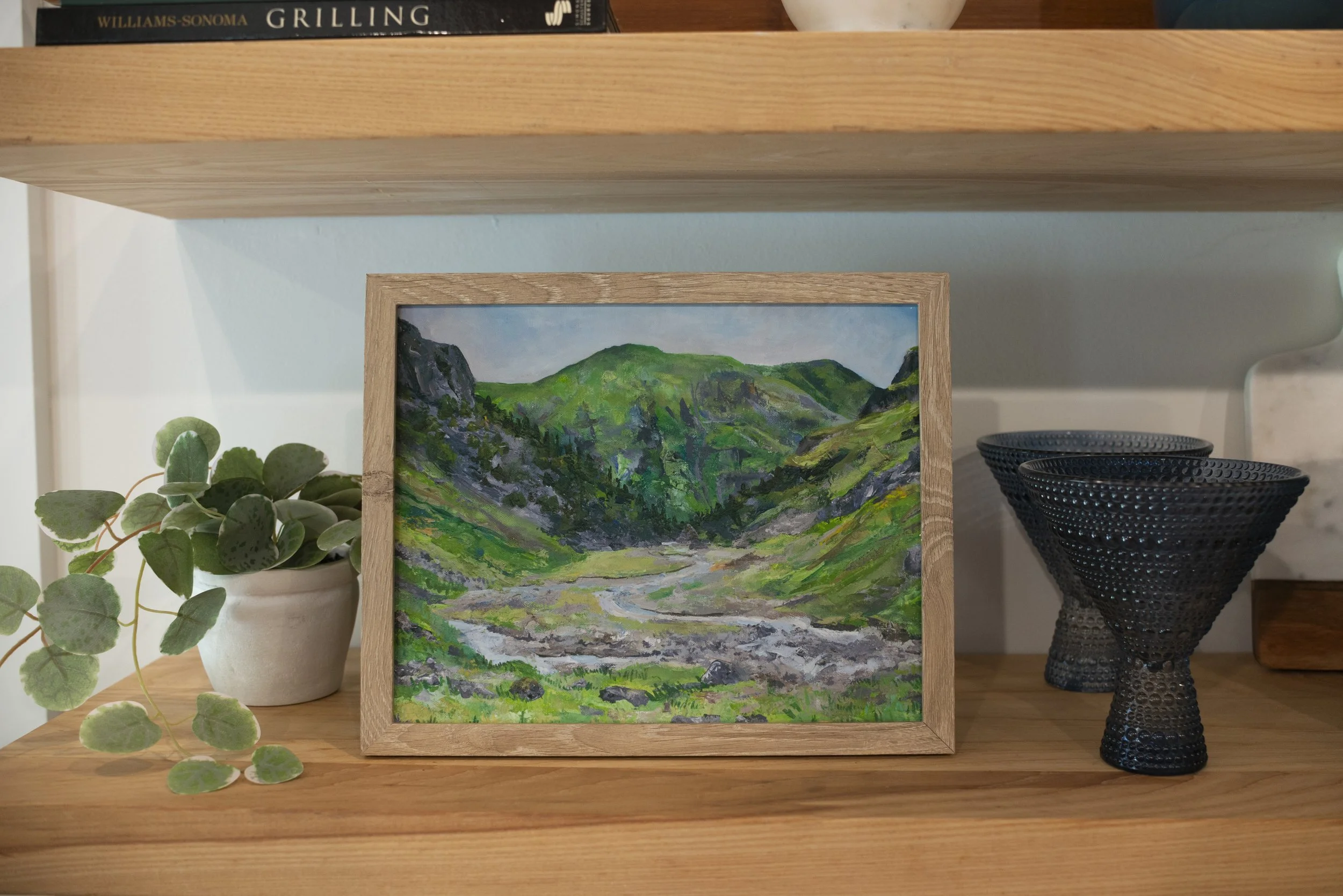 A print of Glencoe in the Scottish Highlands in a wooden frame displayed on a cozy shelf with decorative glasses and an indoor plant.