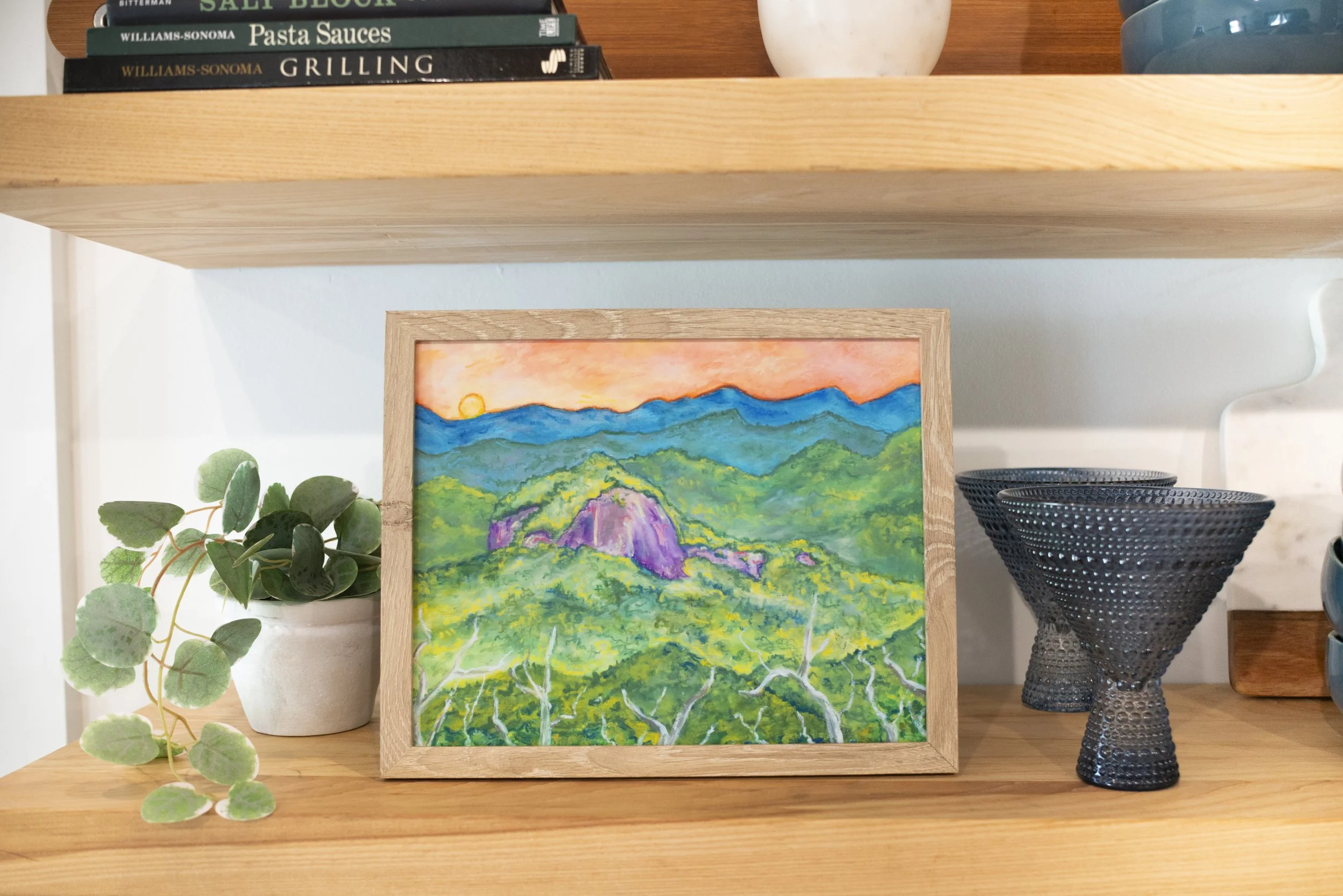 A framed print of a rock monolith surrounded by mountains displayed on a shelf with decorative vases, glasses, dish ware, and cookbooks.