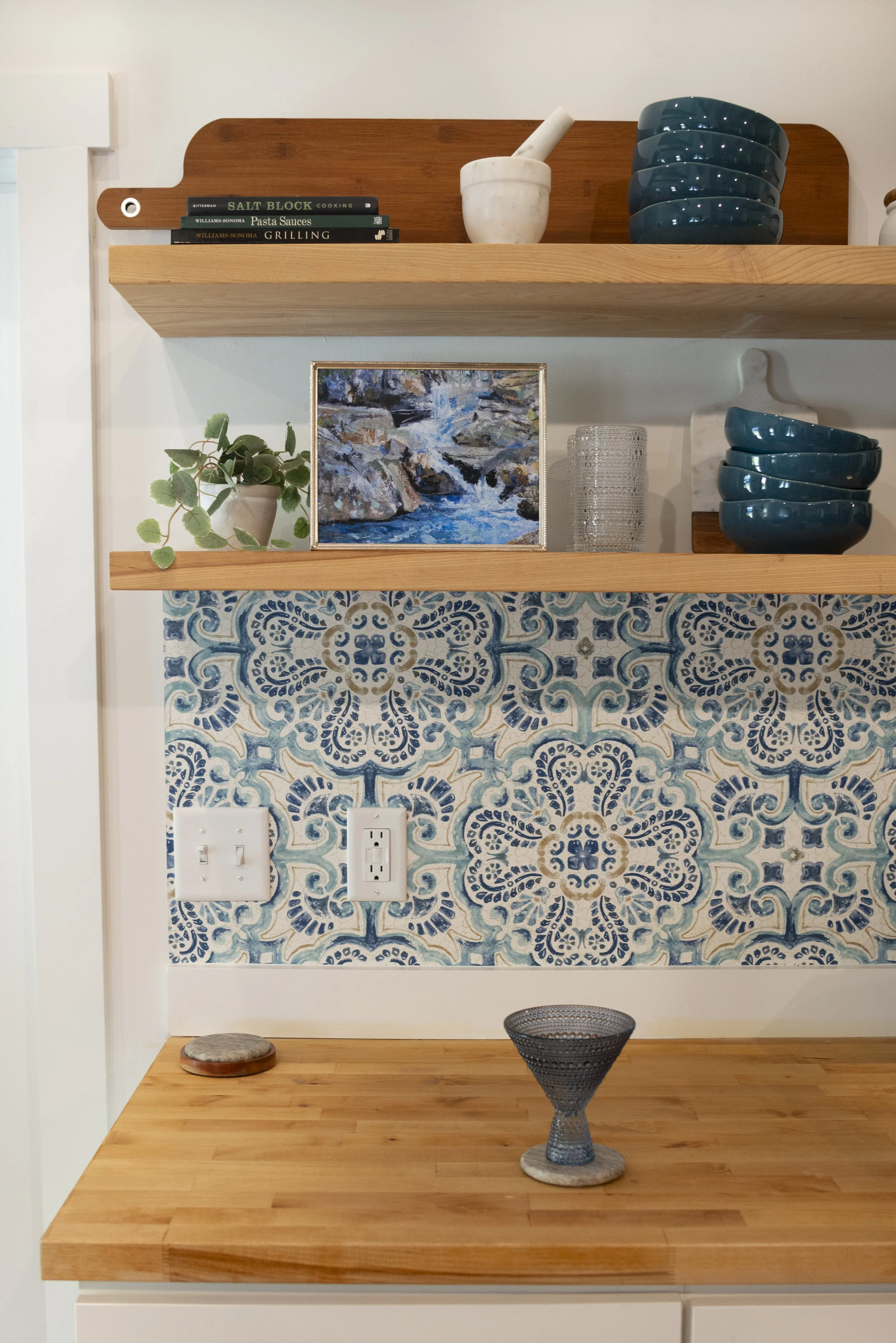 A framed print of a Western NC waterfall, displayed on a kitchen shelf in a cozy, modern kitchen with wooden shelves and colorful accents.