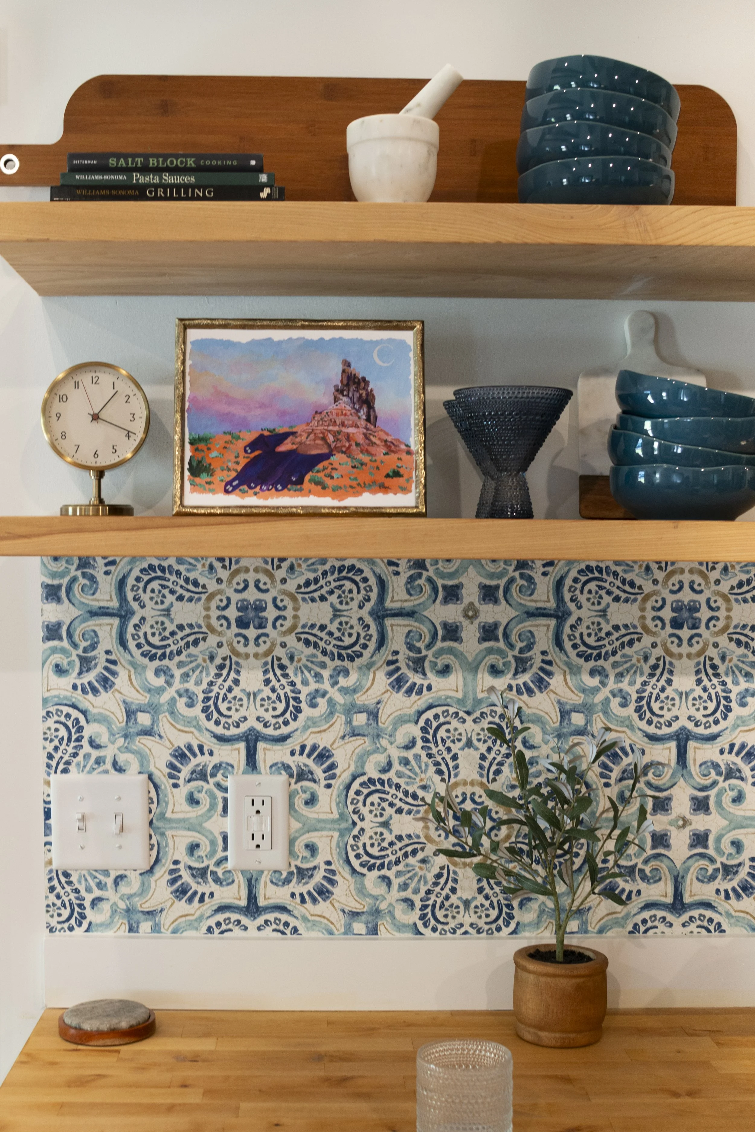 A framed print of a desert scene, displayed on a kitchen shelf in a cozy, modern kitchen with wooden shelves and colorful accents.
