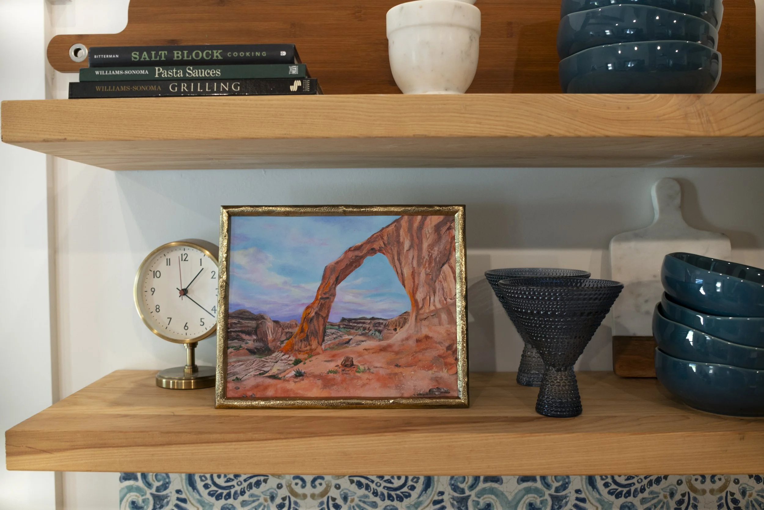 A print of Corona Arch in Moab, UT in a gold frame displayed on a cozy shelf with blue glassware and decorative gold clock.
