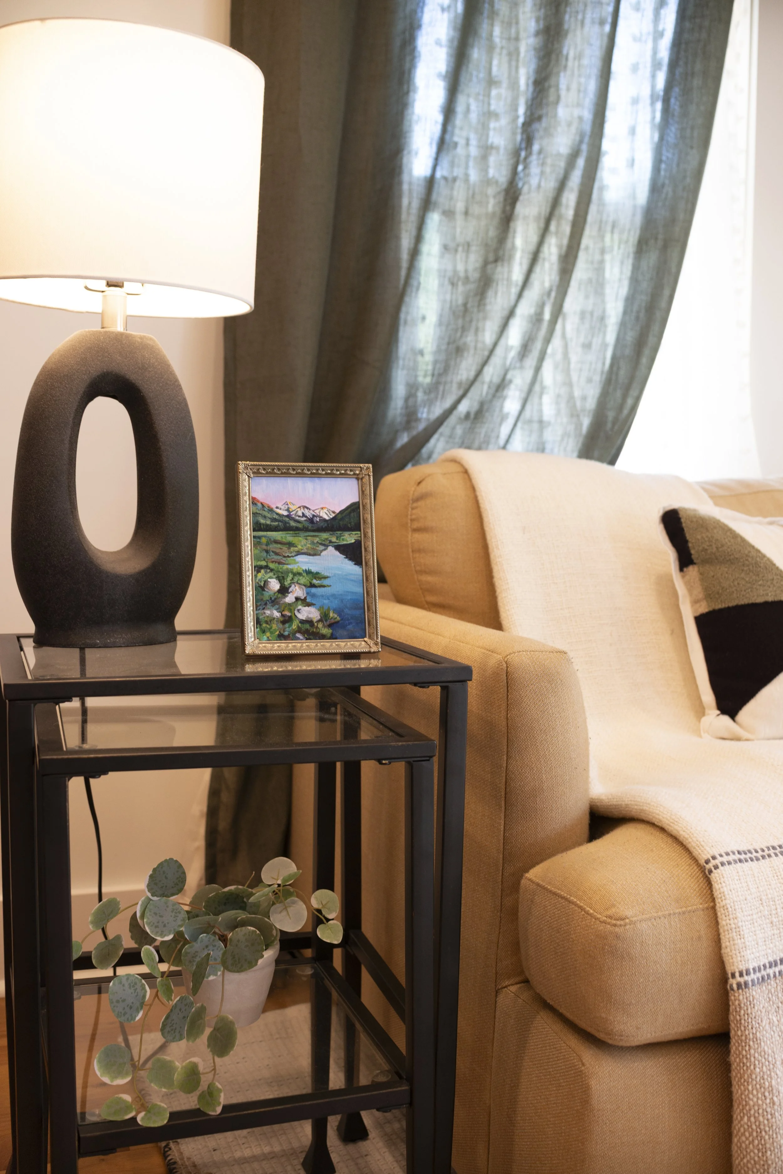 Framed mountain landscape print on a table in a cozy living room, styled with a lamp, indoor plant, and a couch with throw blanket and pillows.